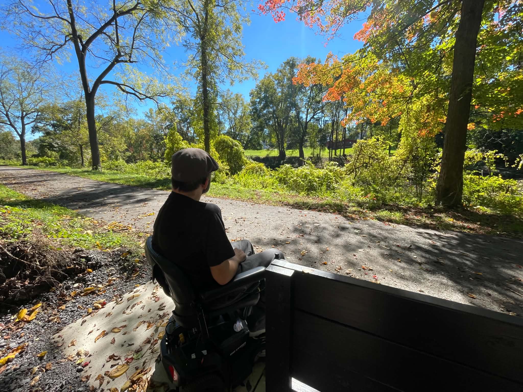 A man using a power wheelchair pauses beside a bench along Newark’s Erie Canalway Trail, looking toward the canal amid sunlit trees and autumn foliage. A man using a power wheelchair pauses beside a bench along Newark’s Erie Canalway Trail, looking toward the canal amid sunlit trees and autumn foliage.