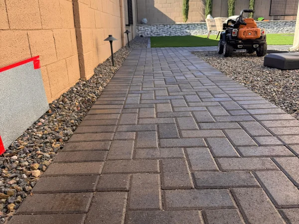 A backyard paver walkway with a light brown wall, gravel, and a child's ride-on toy in the distance.