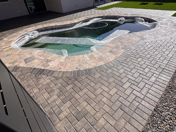 A custom-shaped swimming pool with a bubble cover in place, surrounded by herringbone pattern pavers and gravel on a sunny day.