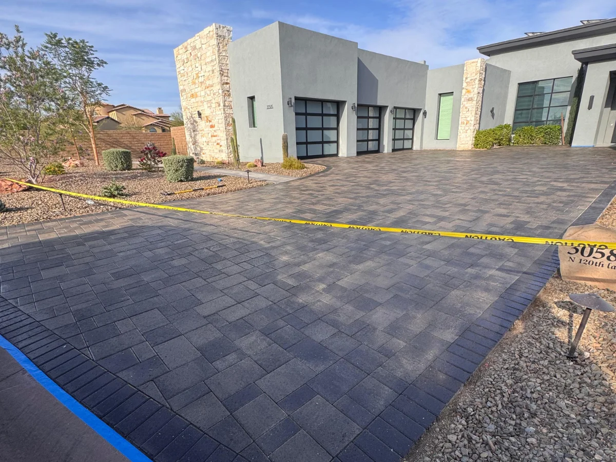 A newly installed paver driveway with a dark border, leading to a modern gray house with garage doors, secured by yellow caution tape.