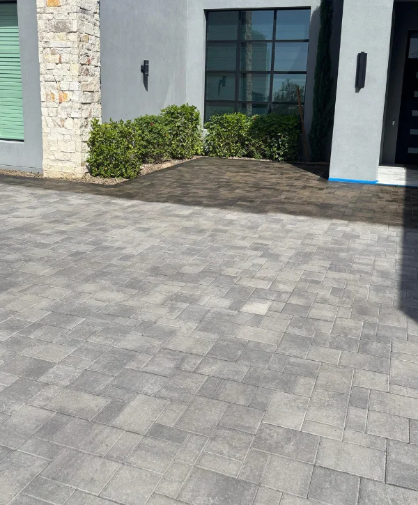 A new patio paver driveway in a modern pattern extends towards a gray building with green bushes.