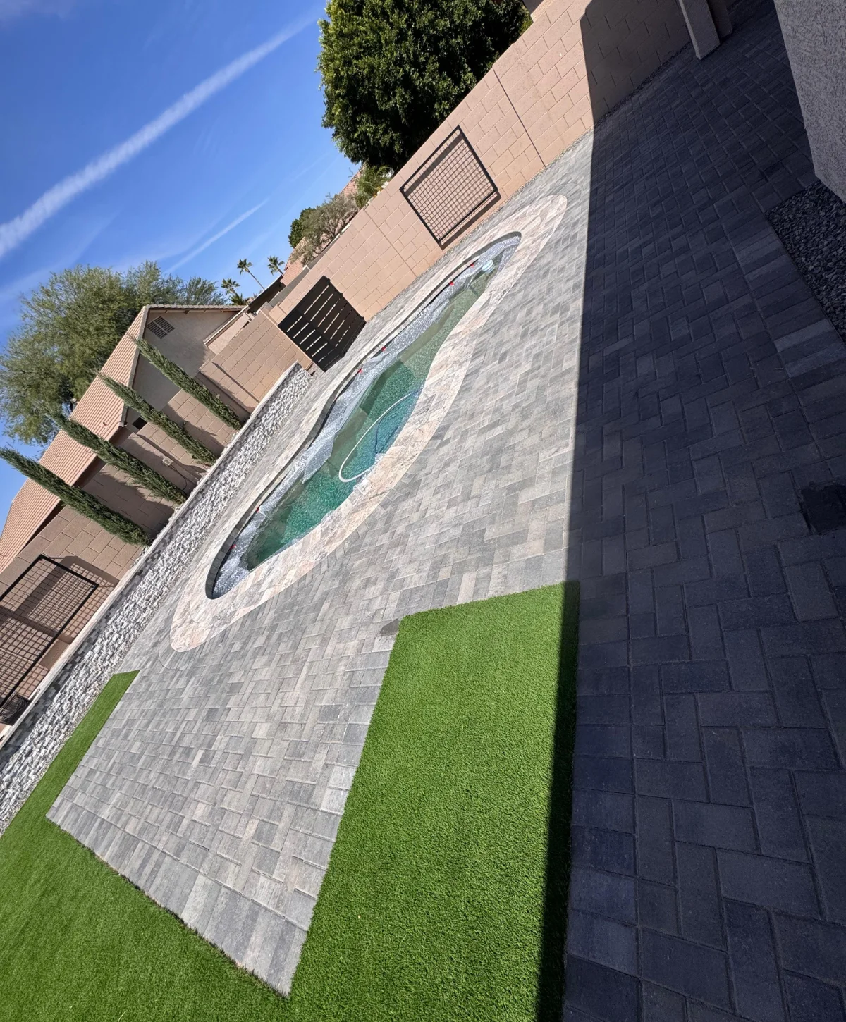 A backyard with a kidney-shaped pool, artificial grass, and two-tone paver patio under a clear blue sky.