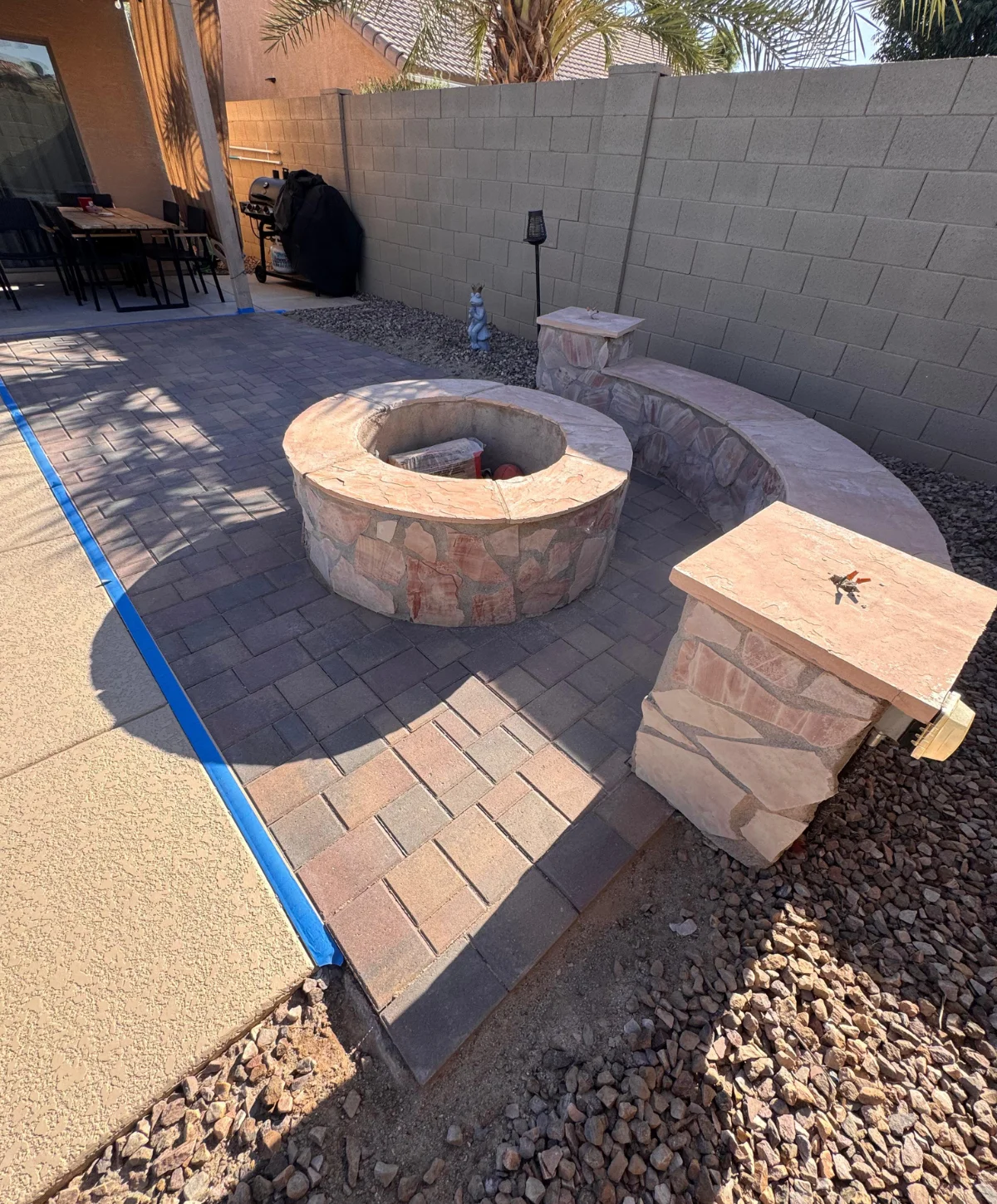 A new backyard paver patio featuring a round stone fire pit, a curved built-in stone bench, and an adjacent stone pillar in a residential outdoor living space.