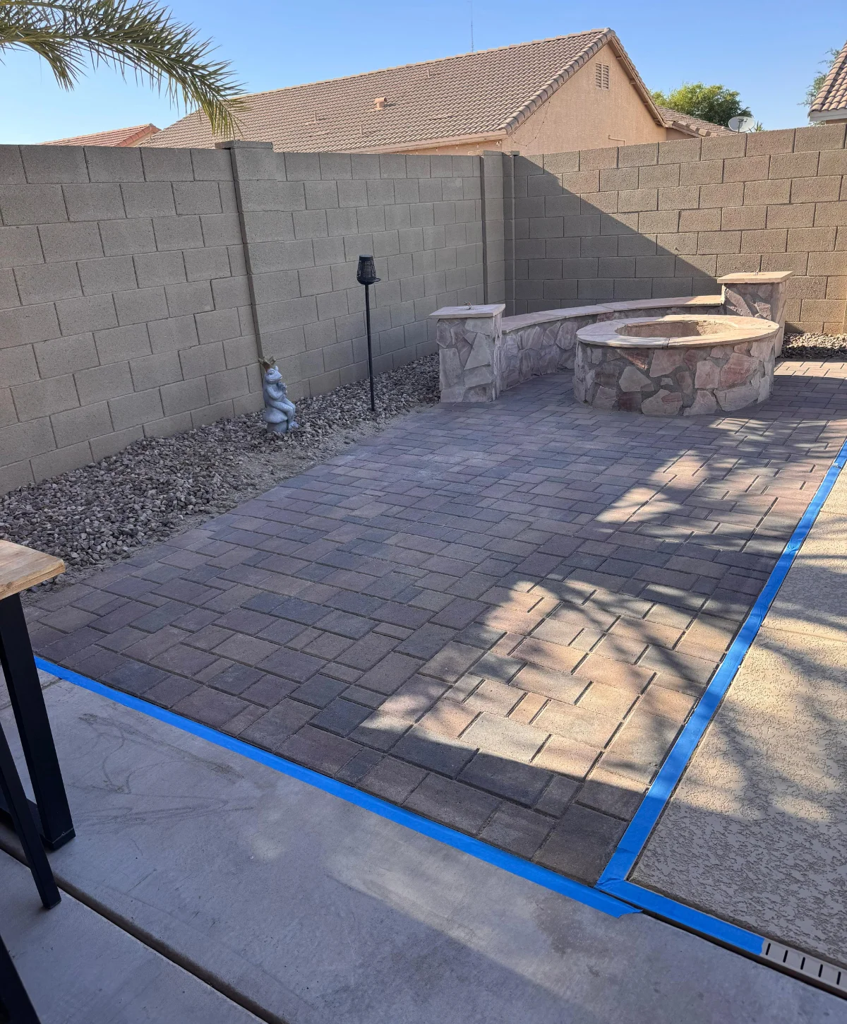 A backyard paver patio and rock-faced fire pit with matching seating surrounded by a block wall and blue tape on the concrete edging.