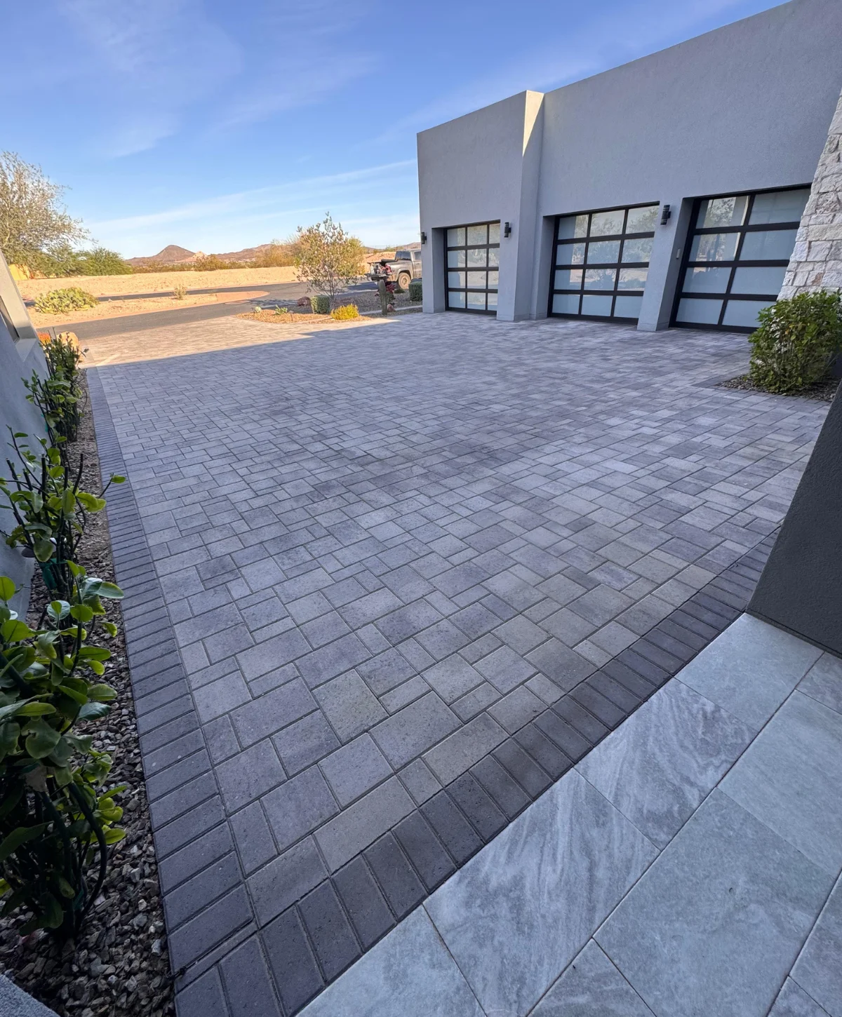 A modern home with a large driveway made of dark gray interlocking pavers, bordered by decorative gravel landscaping. The house features sleek gray exterior walls, black framed garage doors, large windows, and minimalist desert-style landscaping under a sunny sky.