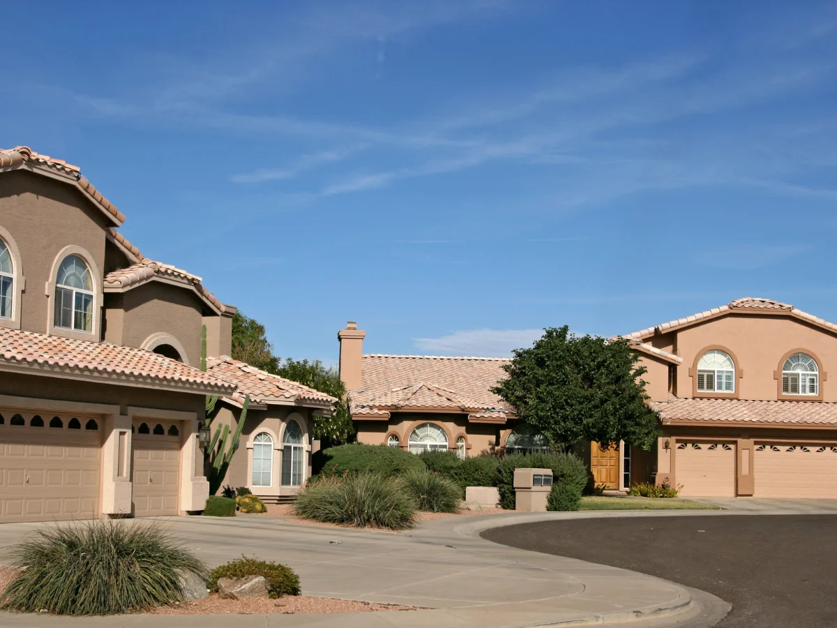 A neighborhood street scene with two large southwestern style homes on a curved driveway under a clear blue sky.