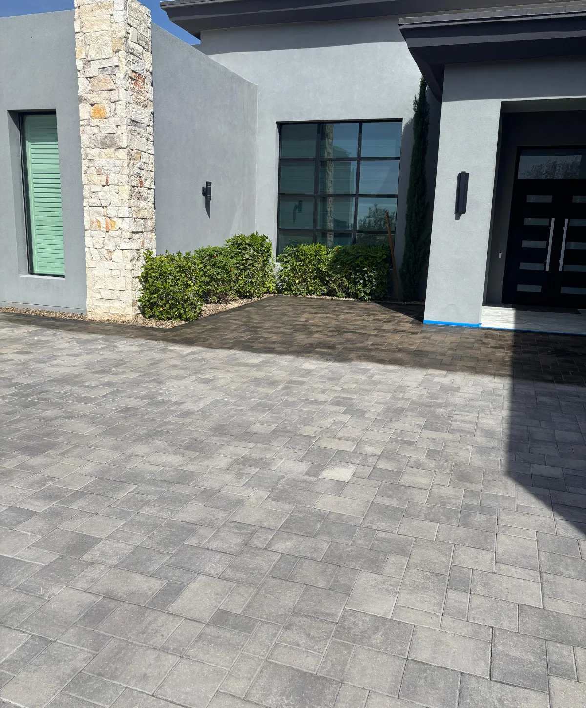 A modern gray house with a stone column and a large paver driveway extending into the foreground.