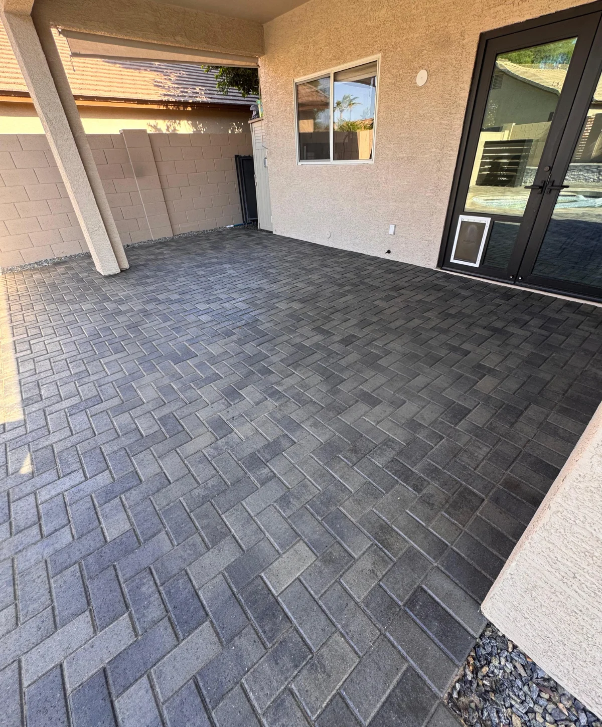 A covered outdoor patio featuring newly installed dark gray herringbone pattern brick pavers leading to a glass door.