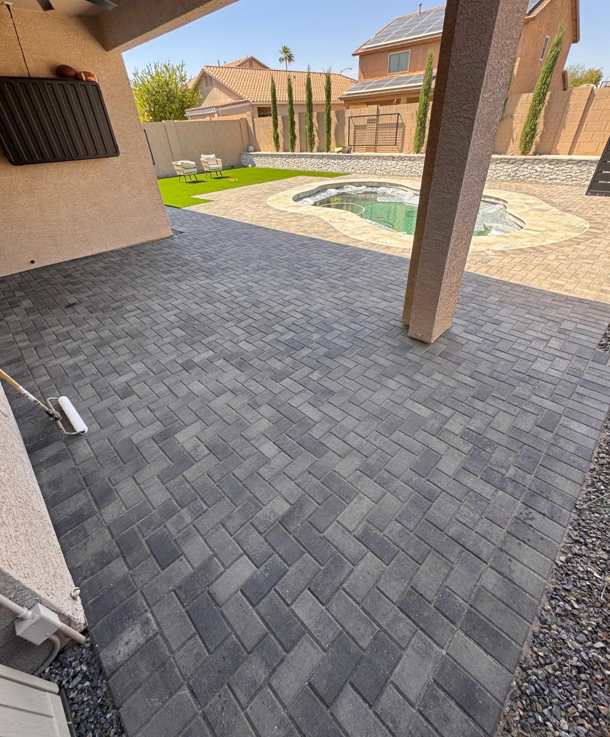 A backyard view showing a newly installed grey paver patio in a diagonal pattern leading to a pool and artificial turf.