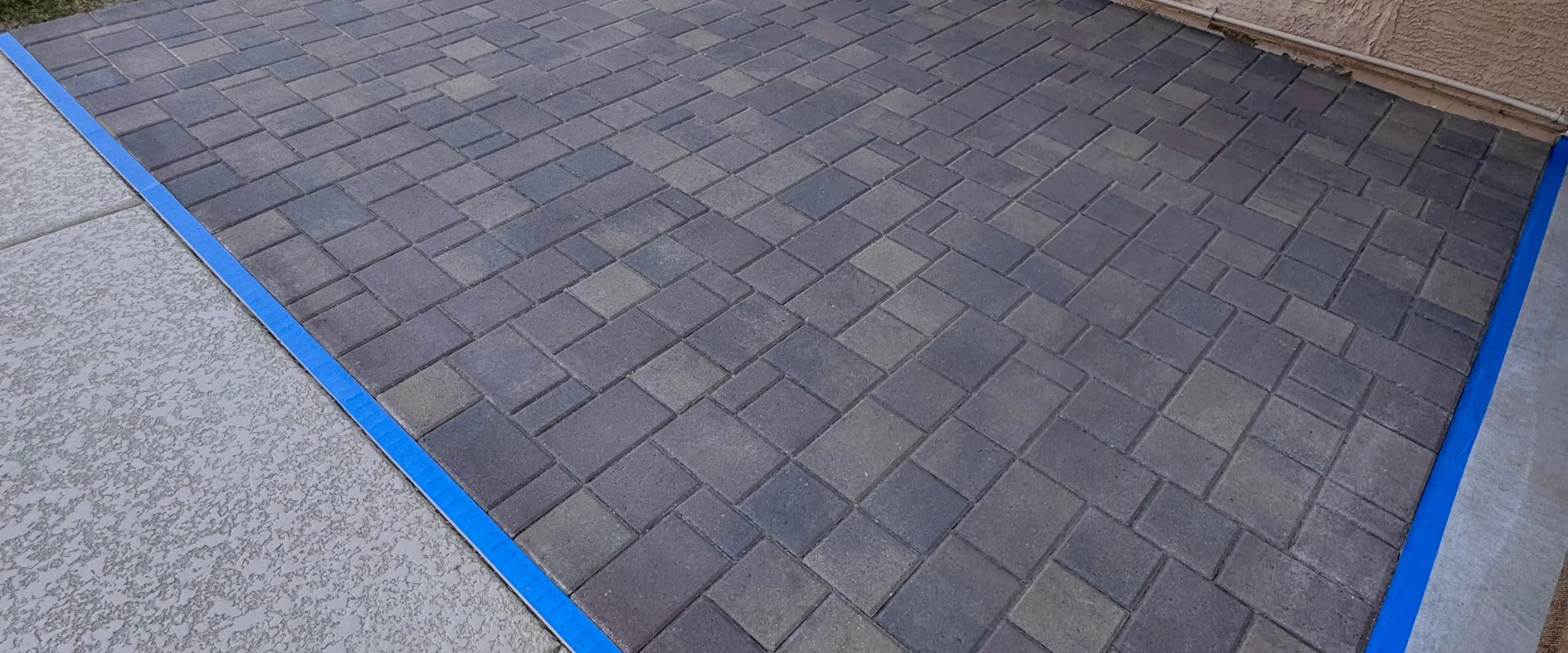 A close-up view of a patterned paver patio bordered by blue painter's tape on two sides.