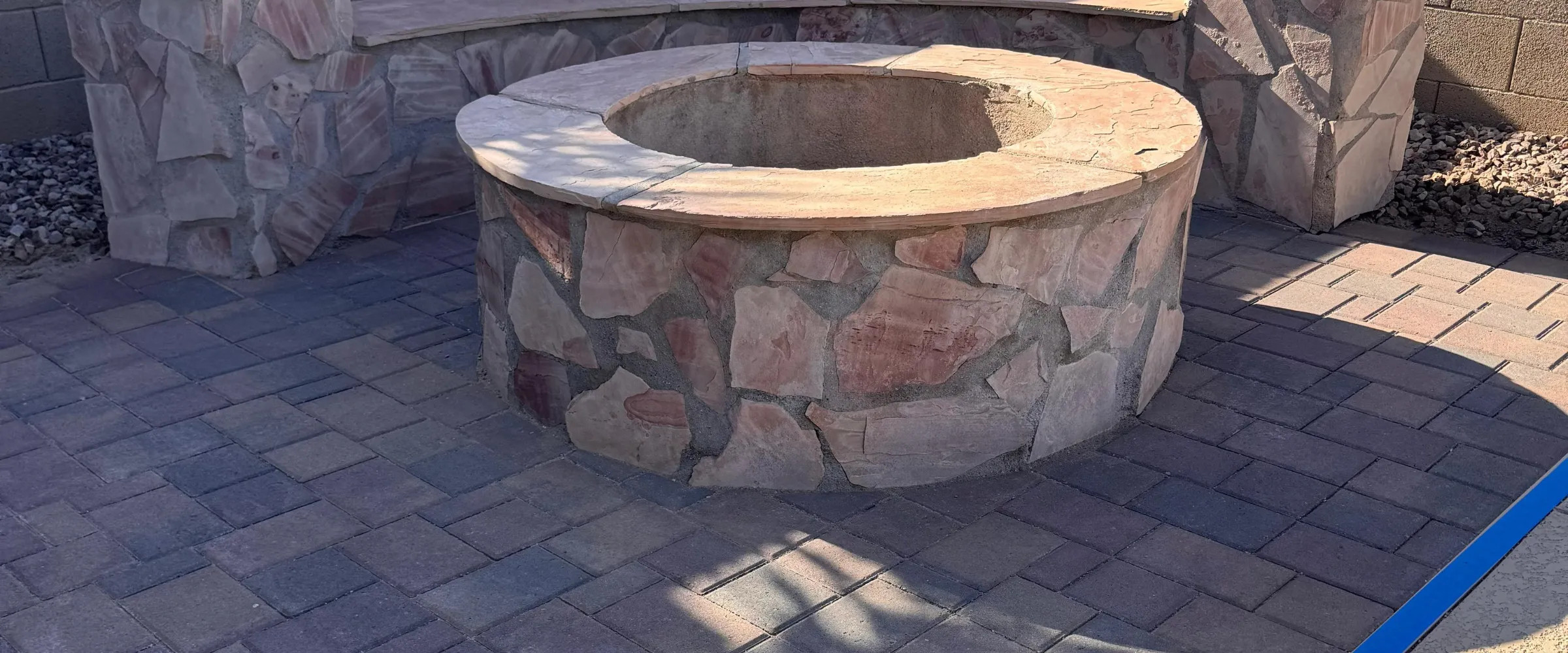 A circular, stacked stone fire pit with a flagstone cap on a paver patio, with a matching stone bench in the background.