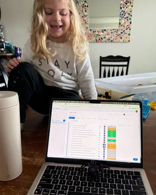 A smiling young girl sitting beside an open laptop with a task board on screen at home