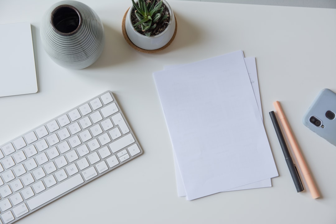 Overhead view of a white desk with keyboard, blank sheets of paper, two pens, a small succulent and a phone.