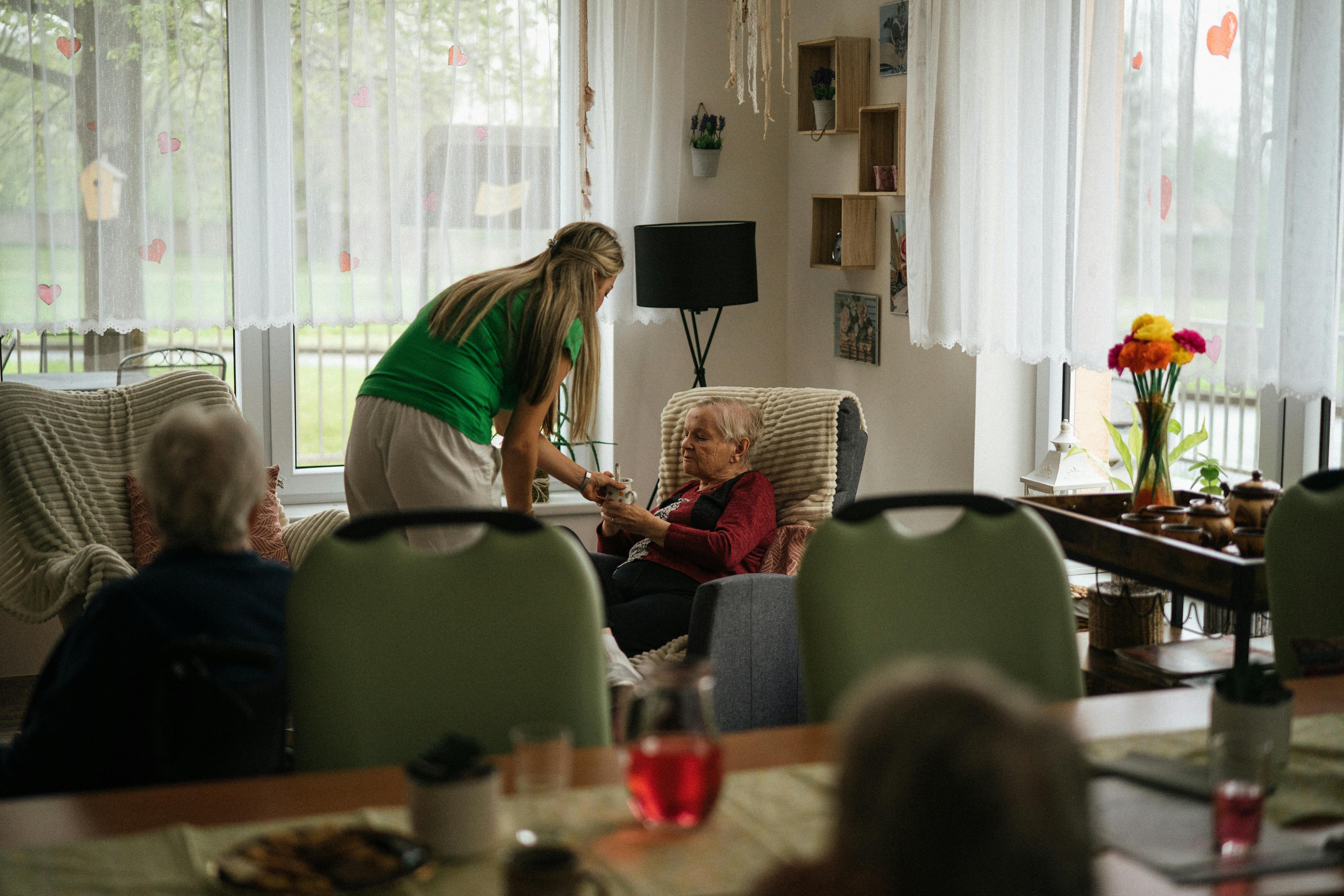 “Caregiver assisting elderly woman with a drink in a cozy living room, senior receiving in-home care and companionship in a comfortable assisted living environment.” “Caregiver assisting elderly woman with a drink in a cozy living room, senior receiving in-home care and companionship in a comfortable assisted living environment.”