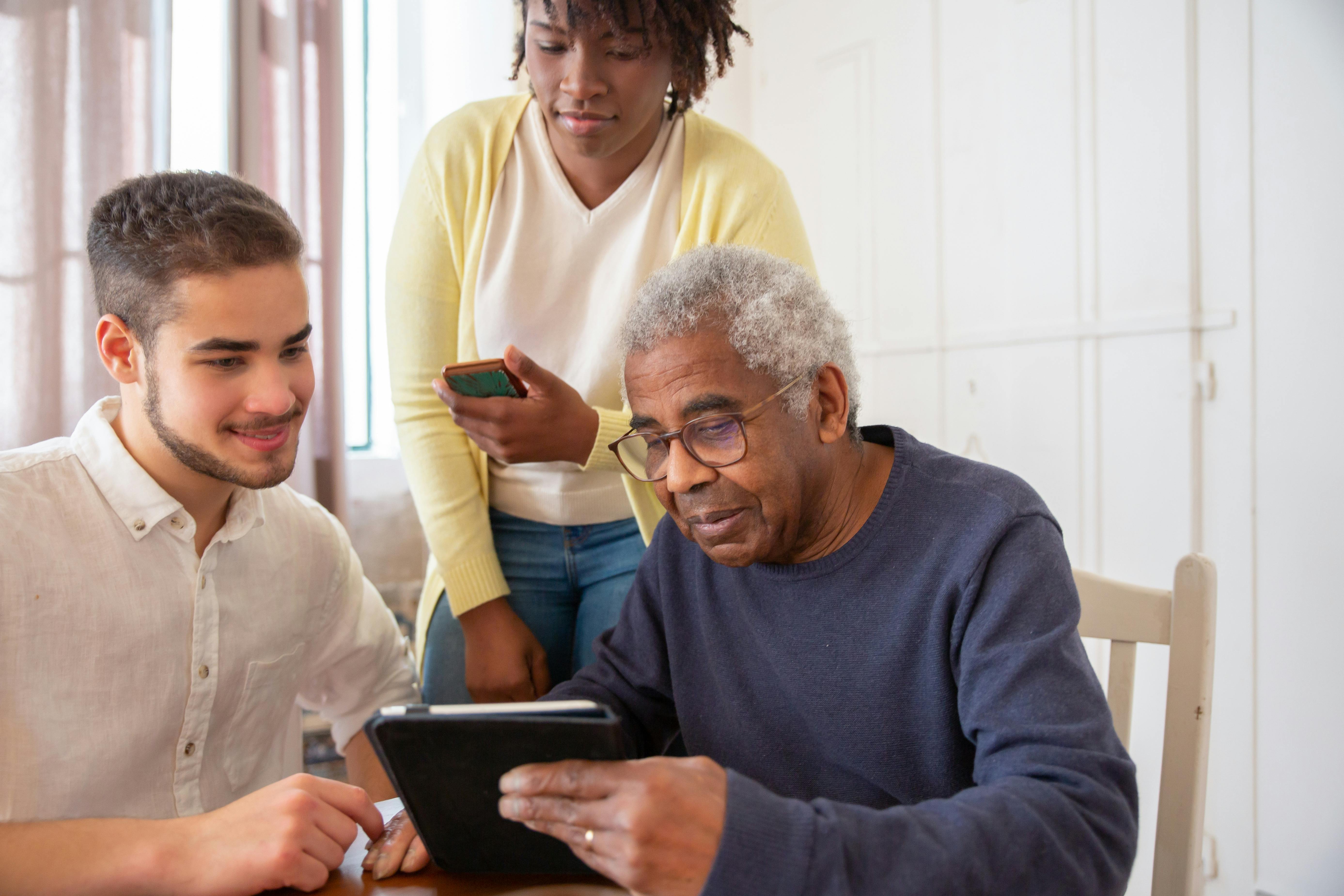 “Caregivers assisting elderly man with tablet technology at home, senior receiving digital support and companionship in a modern in-home care setting.” “Caregivers assisting elderly man with tablet technology at home, senior receiving digital support and companionship in a modern in-home care setting.”