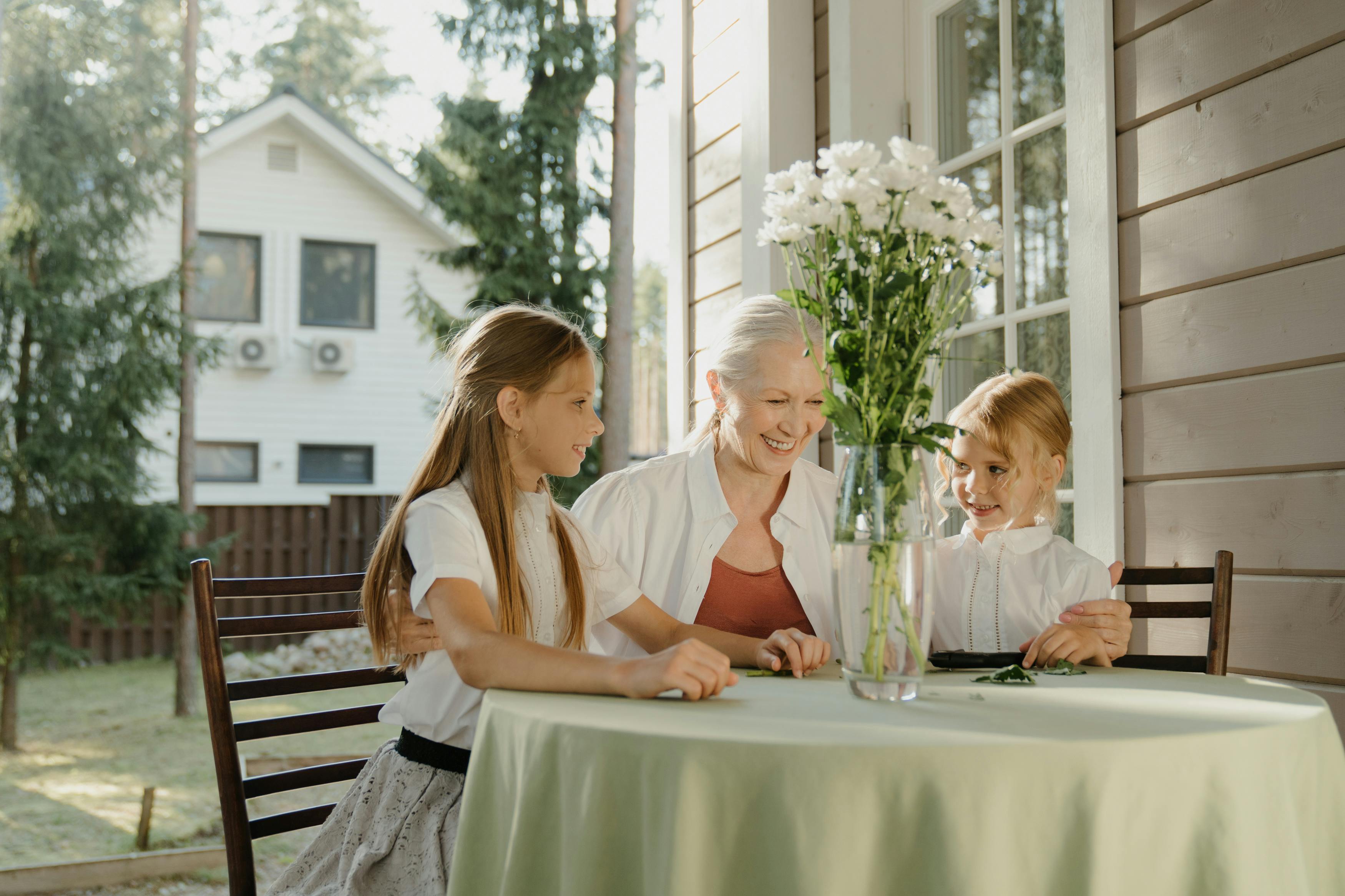 “Grandmother spending quality time with granddaughters at outdoor table, happy multigenerational family bonding in a cozy home setting with natural light.” “Grandmother spending quality time with granddaughters at outdoor table, happy multigenerational family bonding in a cozy home setting with natural light.”