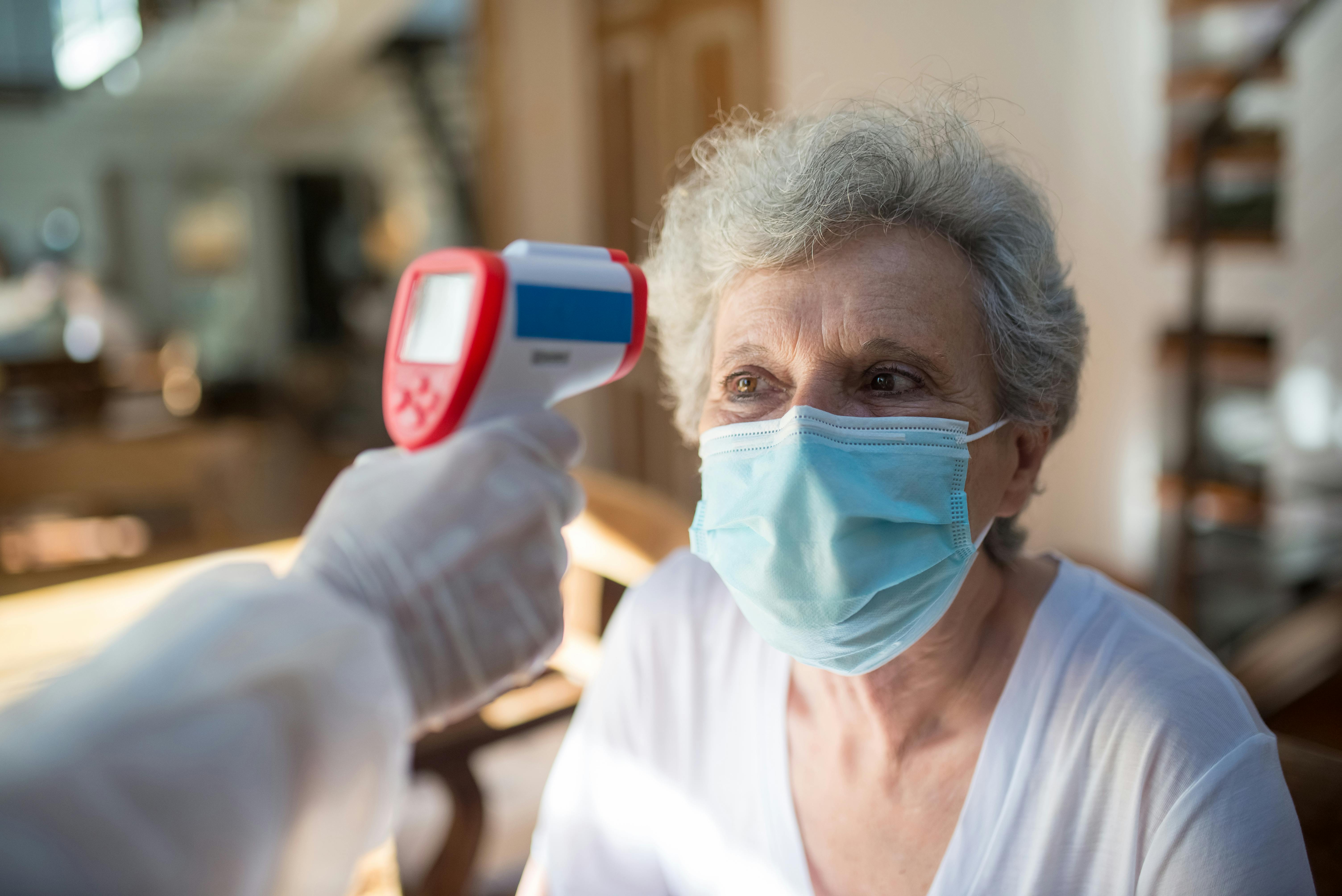 Elderly woman wearing a face mask having her temperature checked with a digital infrared thermometer at home, health safety screening concept Elderly woman wearing a face mask having her temperature checked with a digital infrared thermometer at home, health safety screening concept