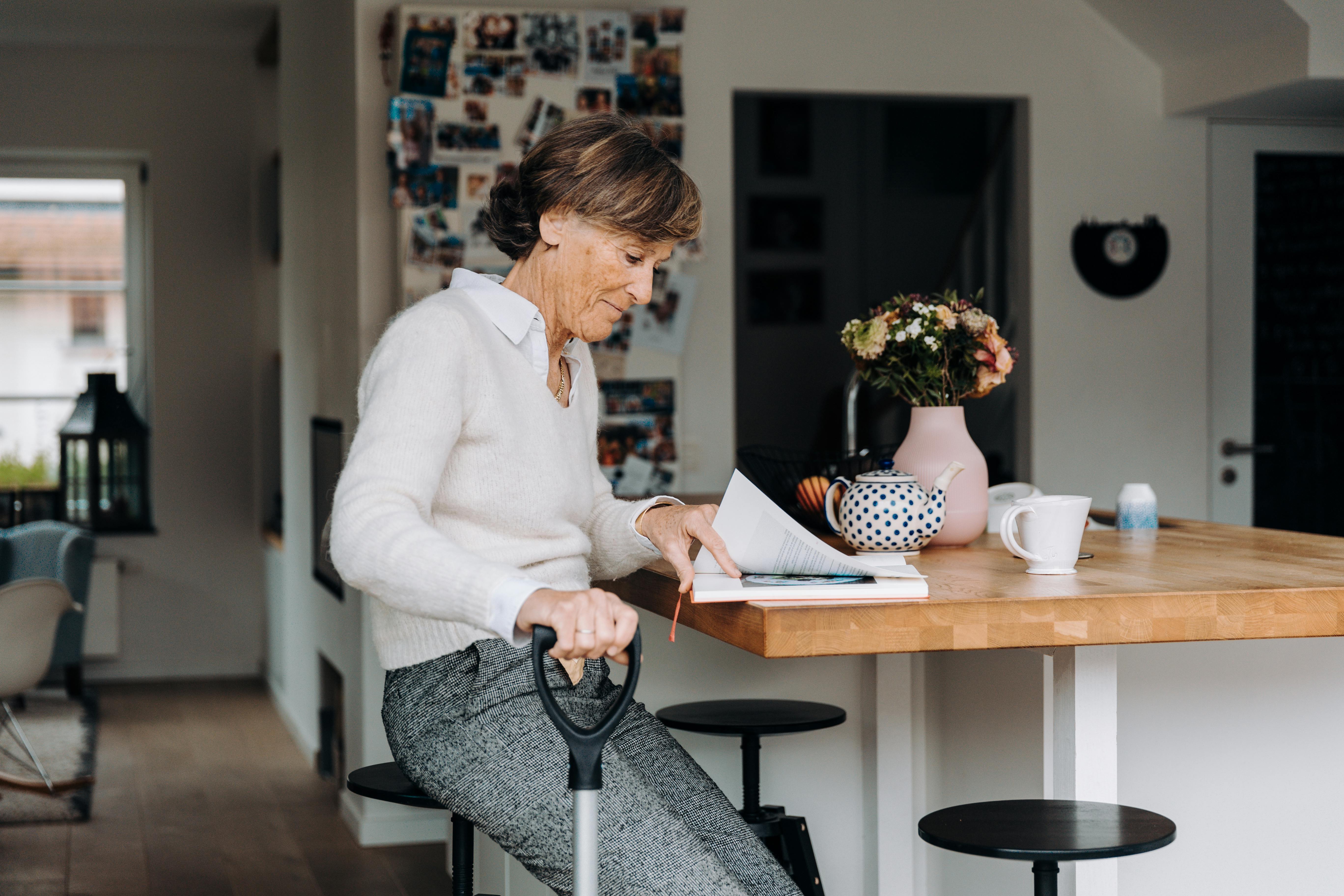 Elderly woman sitting at a kitchen counter using a cane while reading documents at home, representing independent senior living and daily care routines. Elderly woman sitting at a kitchen counter using a cane while reading documents at home, representing independent senior living and daily care routines.