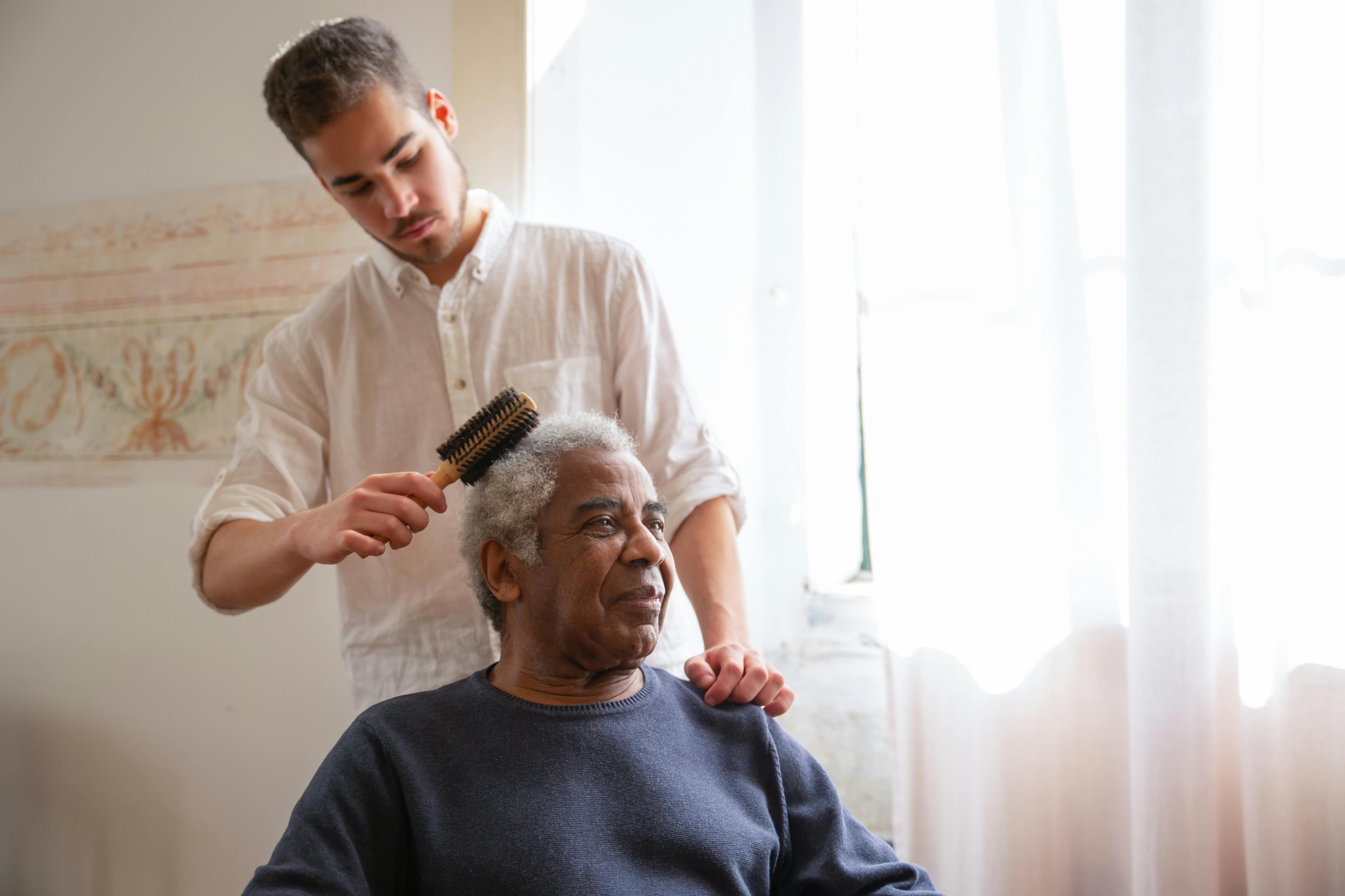 Young male caregiver brushing the hair of an elderly man in a brightly lit room. Young male caregiver brushing the hair of an elderly man in a brightly lit room.