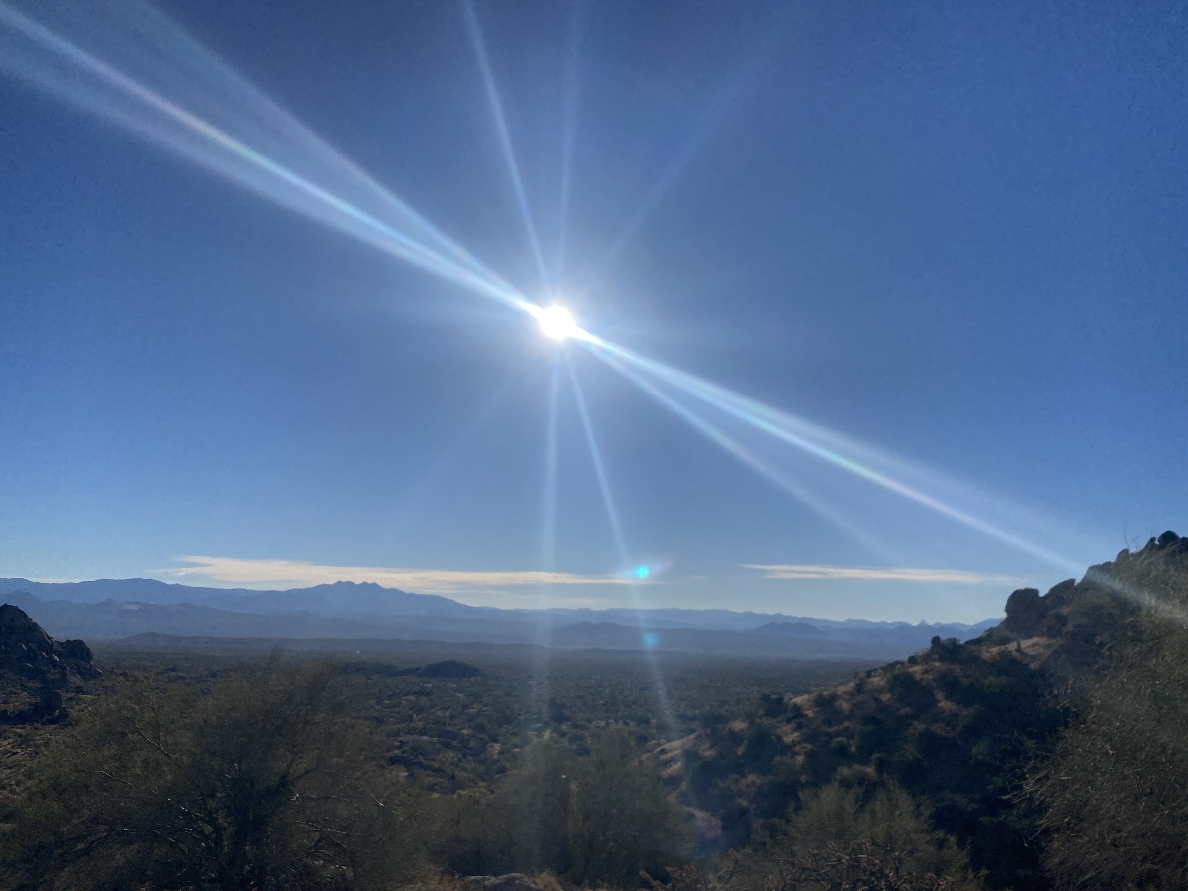 A panoramic view of a vast valley and distant mountains under a clear blue sky with a powerful, bright sunburst radiating light beams.