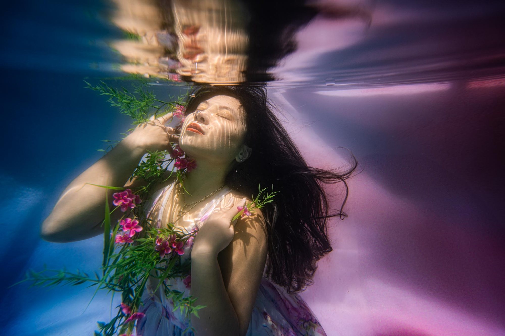 A woman underwater with eyes closed, pink flowers at her shoulder, hair drifting