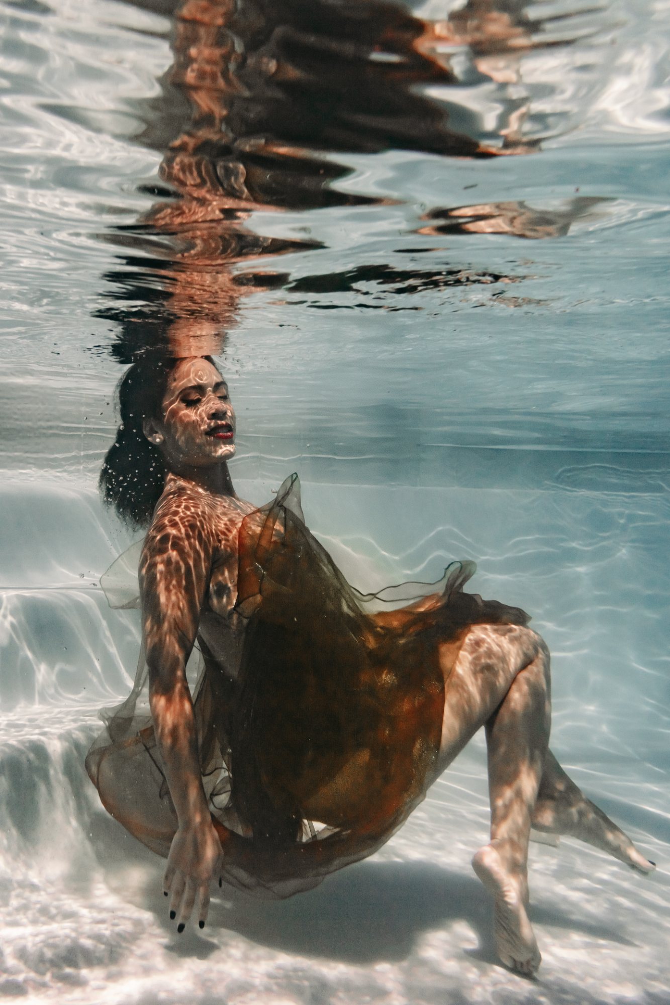 A woman in sheer amber fabric reclining underwater, sunlight rippling across her skin