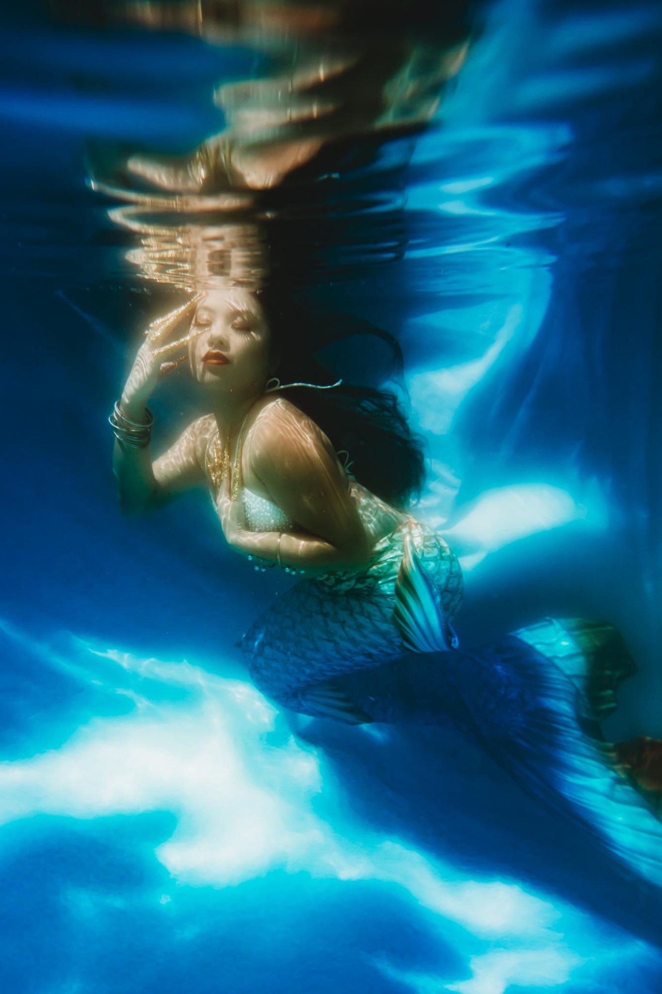 A woman in a mermaid tail drifting in bright blue water, eyes closed