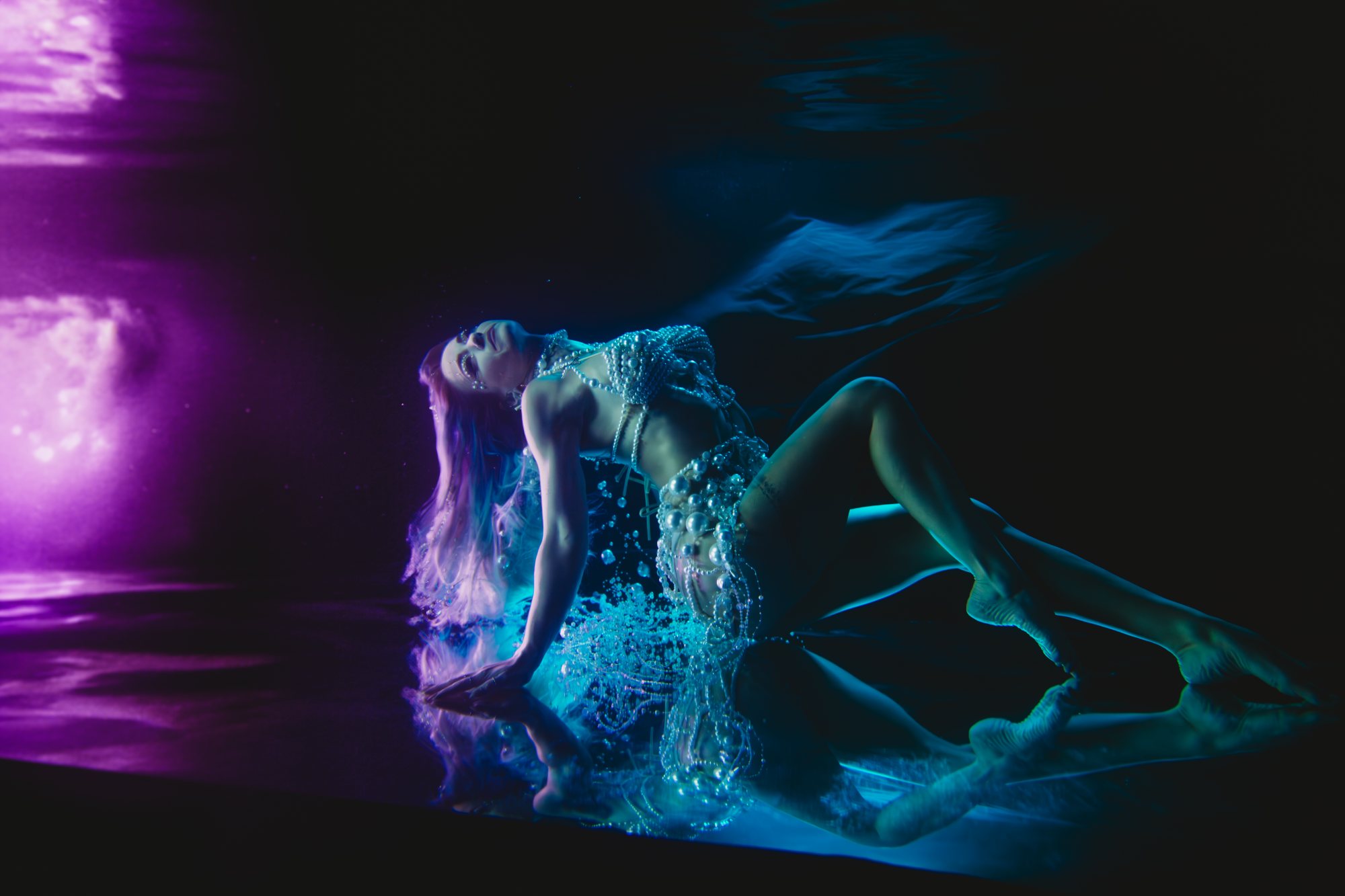 A woman in a pearl bodysuit arched back across a reflective pool floor in violet and teal light