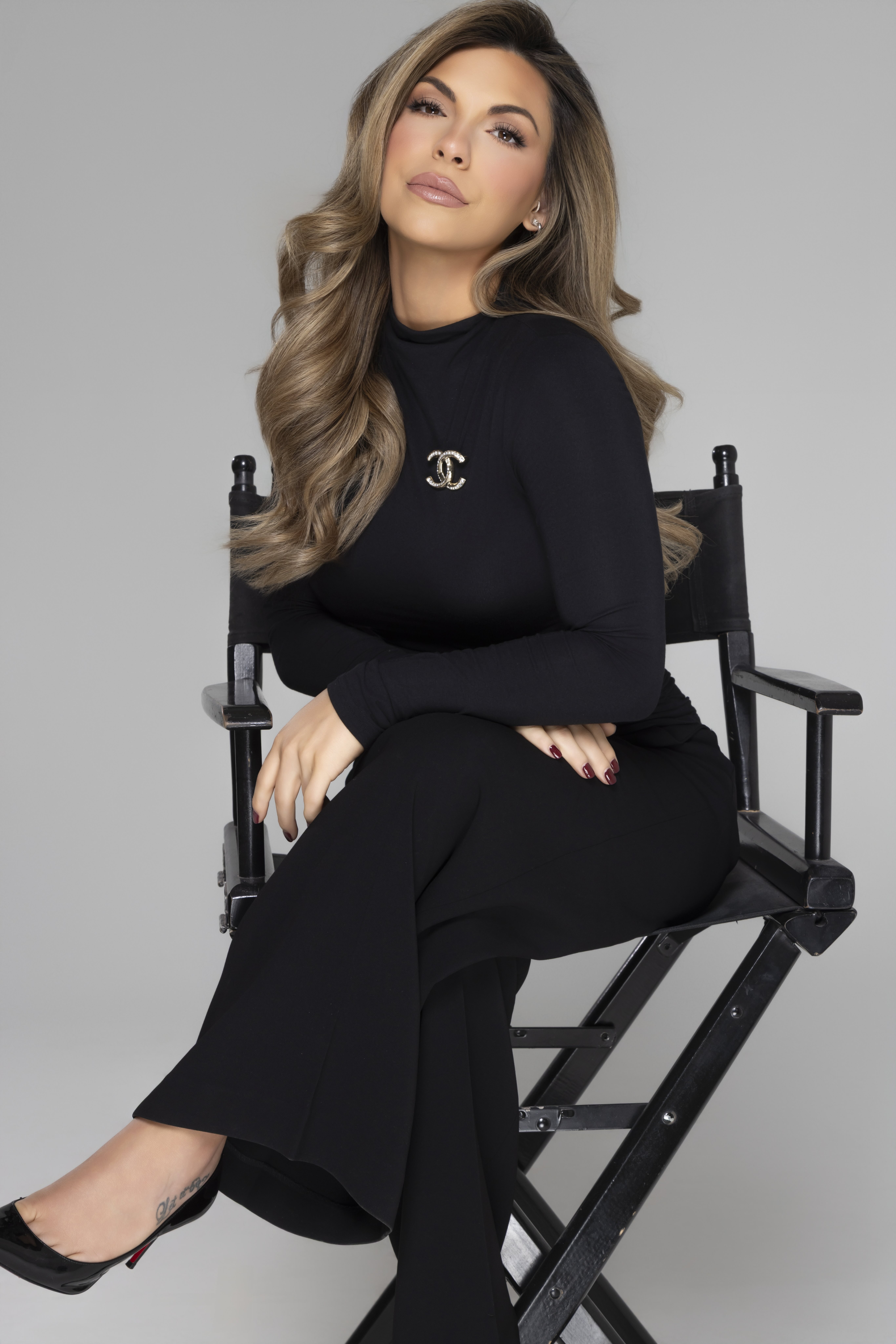 Woman with long wavy blonde hair seated on a black director's chair, wearing a black long-sleeve top with a Chanel brooch and black pants.