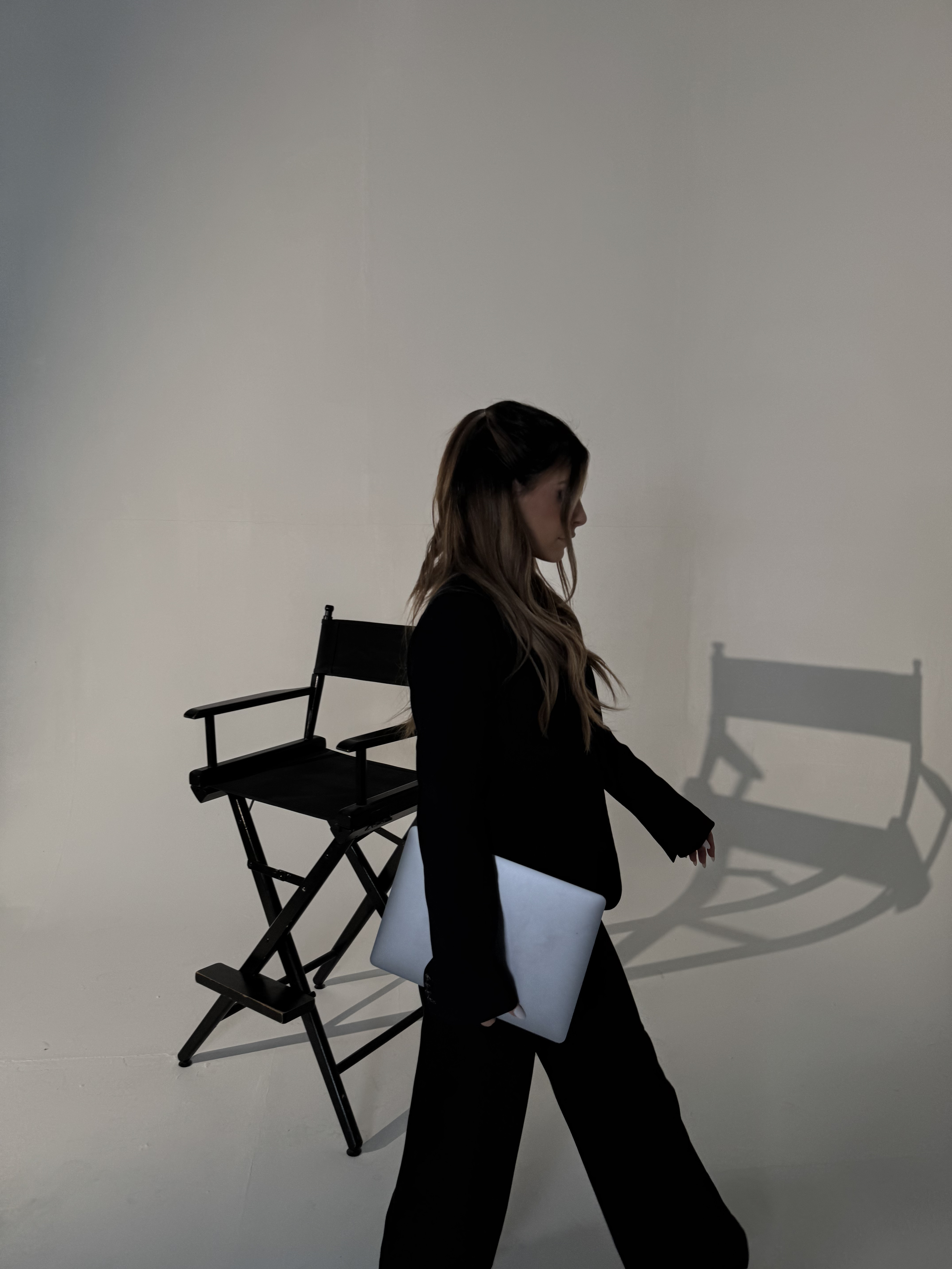 Woman in black outfit holding a silver laptop walking past a black director's chair casting a shadow on a beige wall.