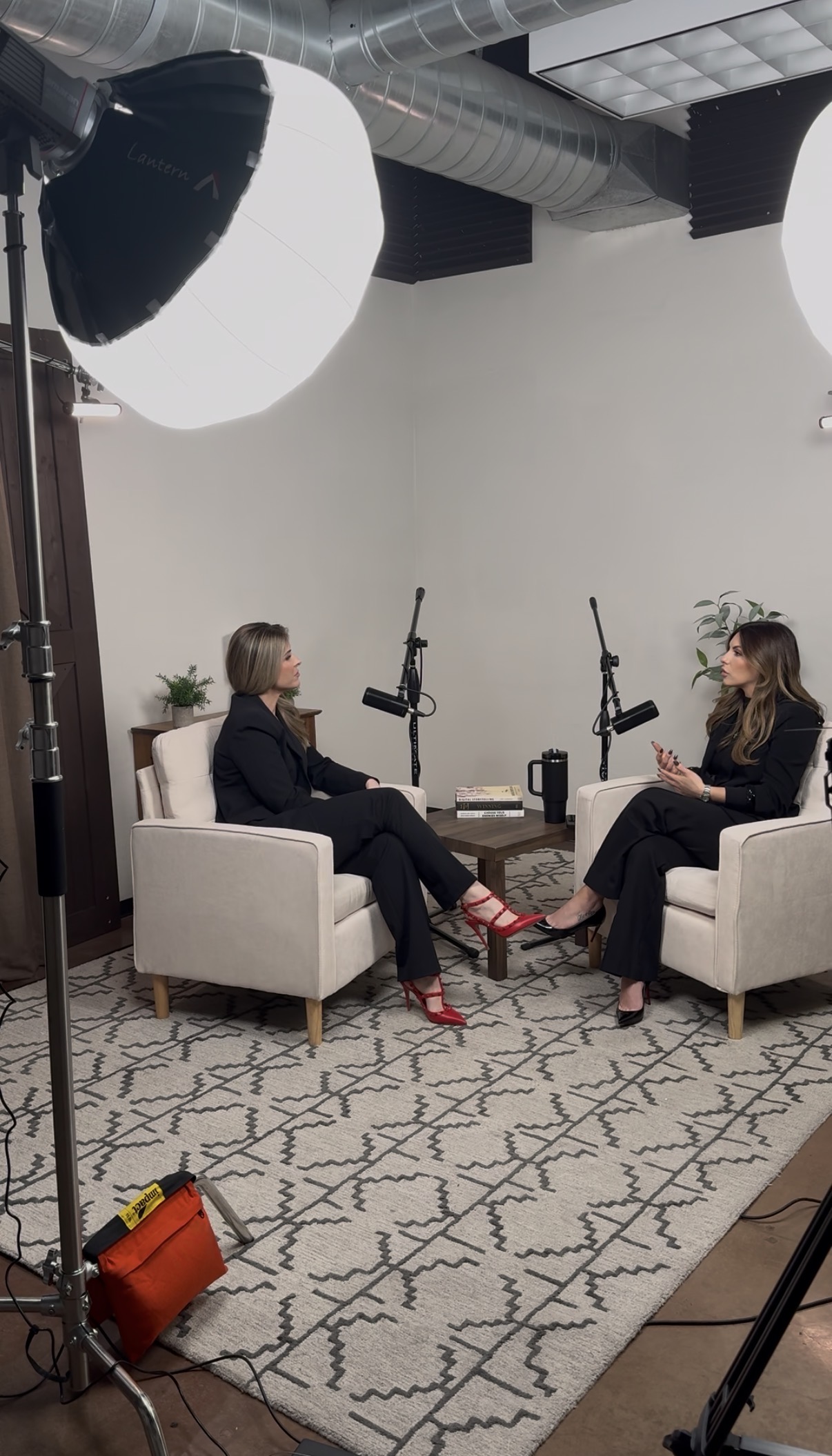 Two women in black suits sitting opposite each other in armchairs, engaged in conversation with microphones between them in a studio setting.