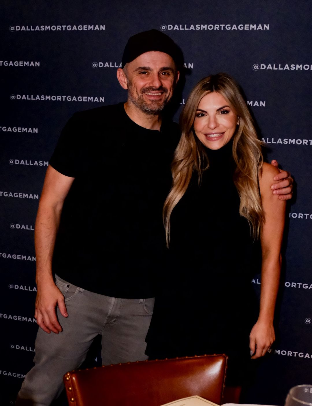 Man in a black cap and t-shirt with arm around smiling woman in a black sleeveless top, standing in front of a backdrop with repeated @DallasMortgageMan text.