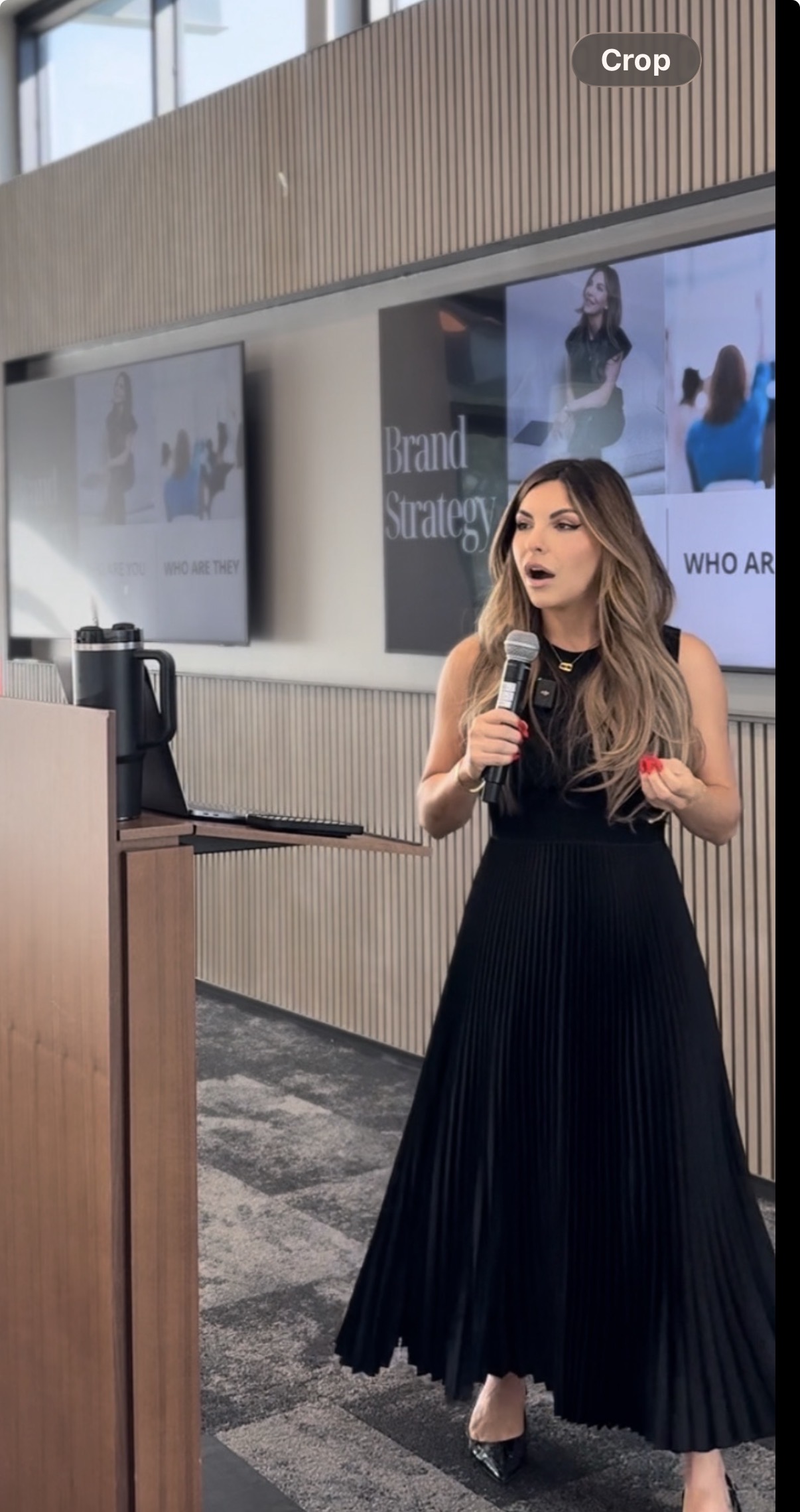 Woman in a black sleeveless dress speaking into a microphone during a presentation next to a podium and a screen displaying 'Brand Strategy'.