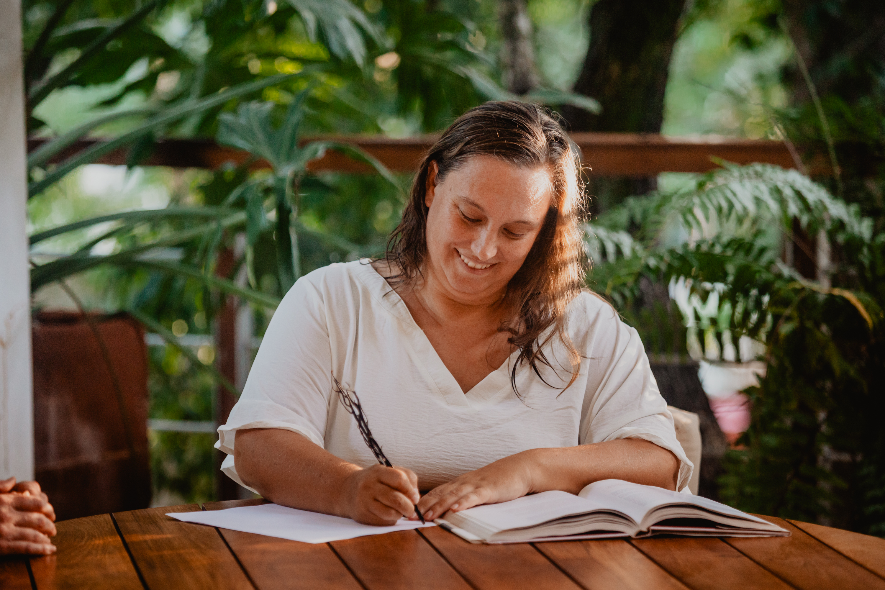 Jaime White sitting at a wooden table outdoors, surrounded by lush greenery. She is smiling and writing in an open notebook, wearing a white blouse with rolled-up sleeves. The atmosphere is calm and focused, with natural light filtering through the trees.