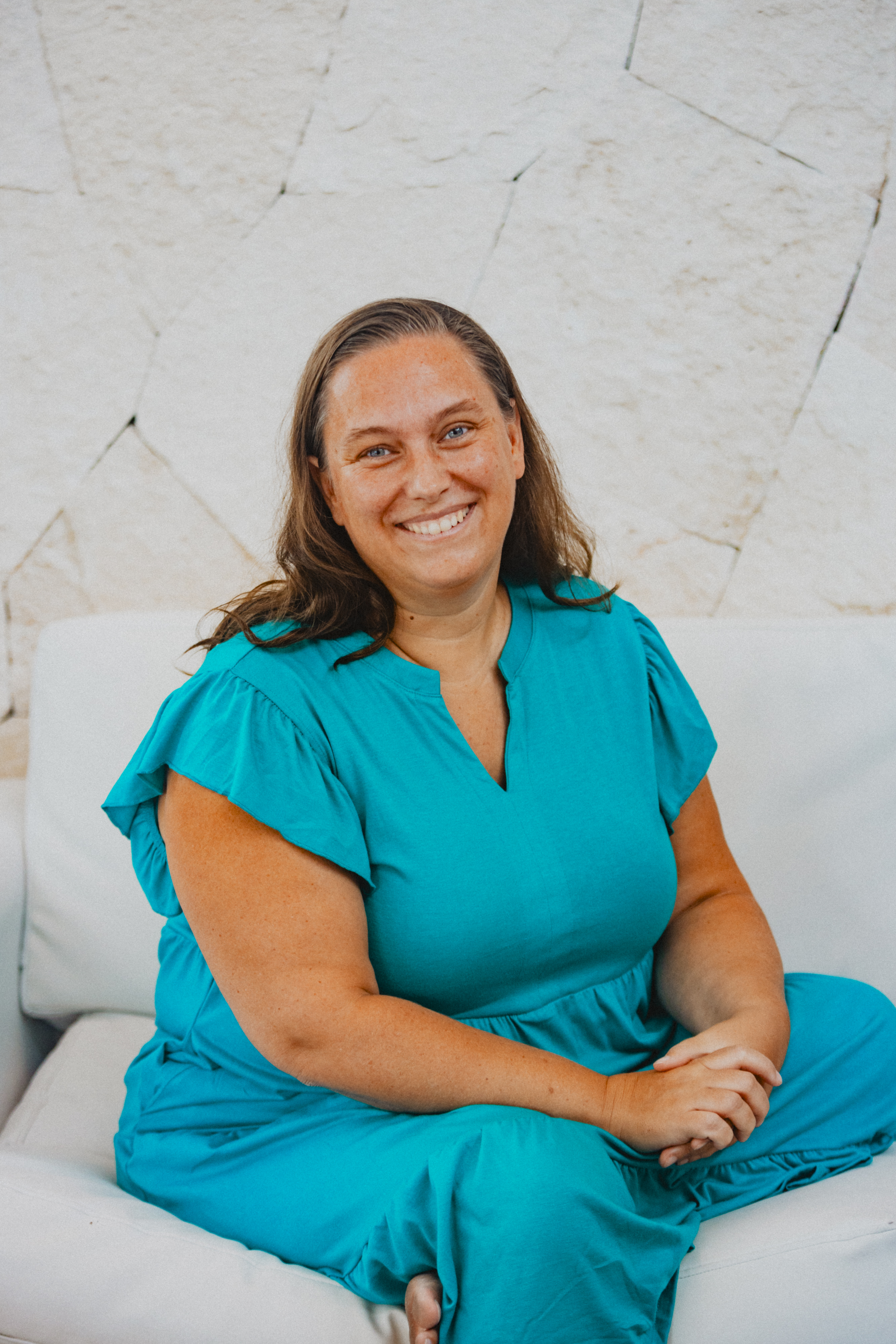 Jaime White sitting on a white couch, smiling warmly at the camera. She is wearing a teal outfit with flutter sleeves, and the background features a textured stone wall. Her hands are gently clasped in her lap, and she exudes a relaxed and friendly presence.