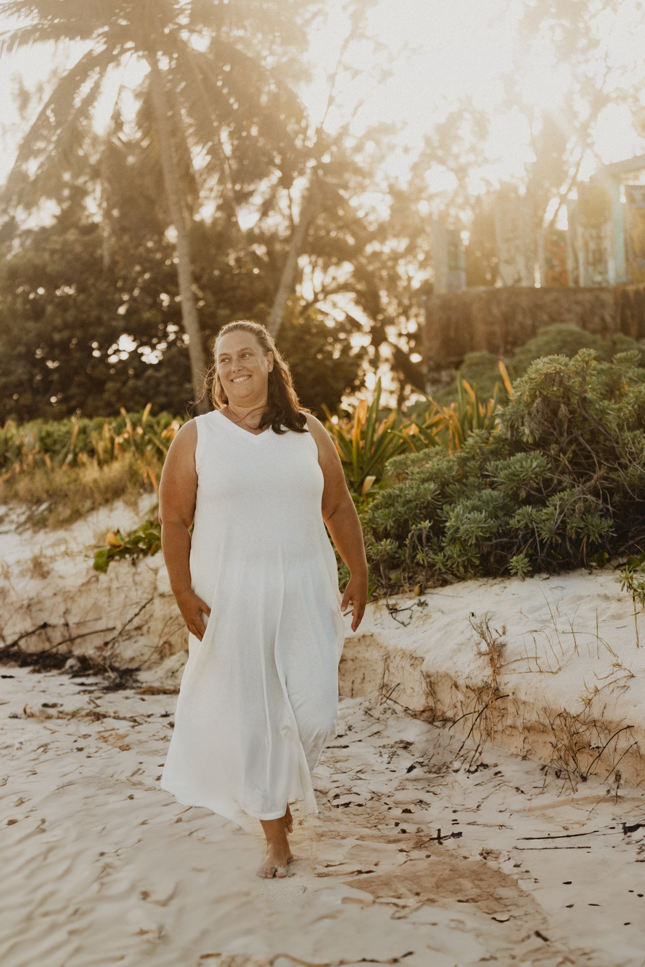 Woman (Jaime White) in a sleeveless white dress walking barefoot along a sandy beach at sunset, surrounded by tropical greenery and palm trees. She is smiling and looking off to the side, with warm golden sunlight filtering through the trees in the background.