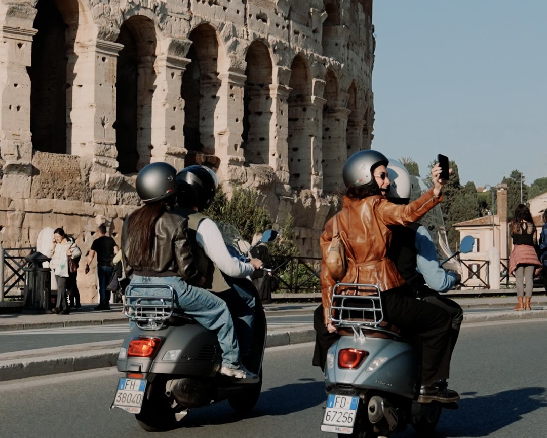 Two Vespas riding past the Colosseum, a passenger taking a selfie