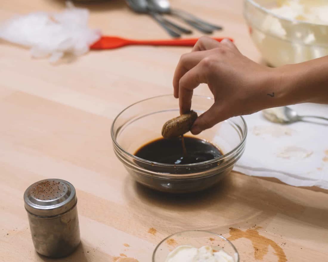 A hand dipping a savoiardo into espresso beside a dusting of cocoa and a glass of tiramisu