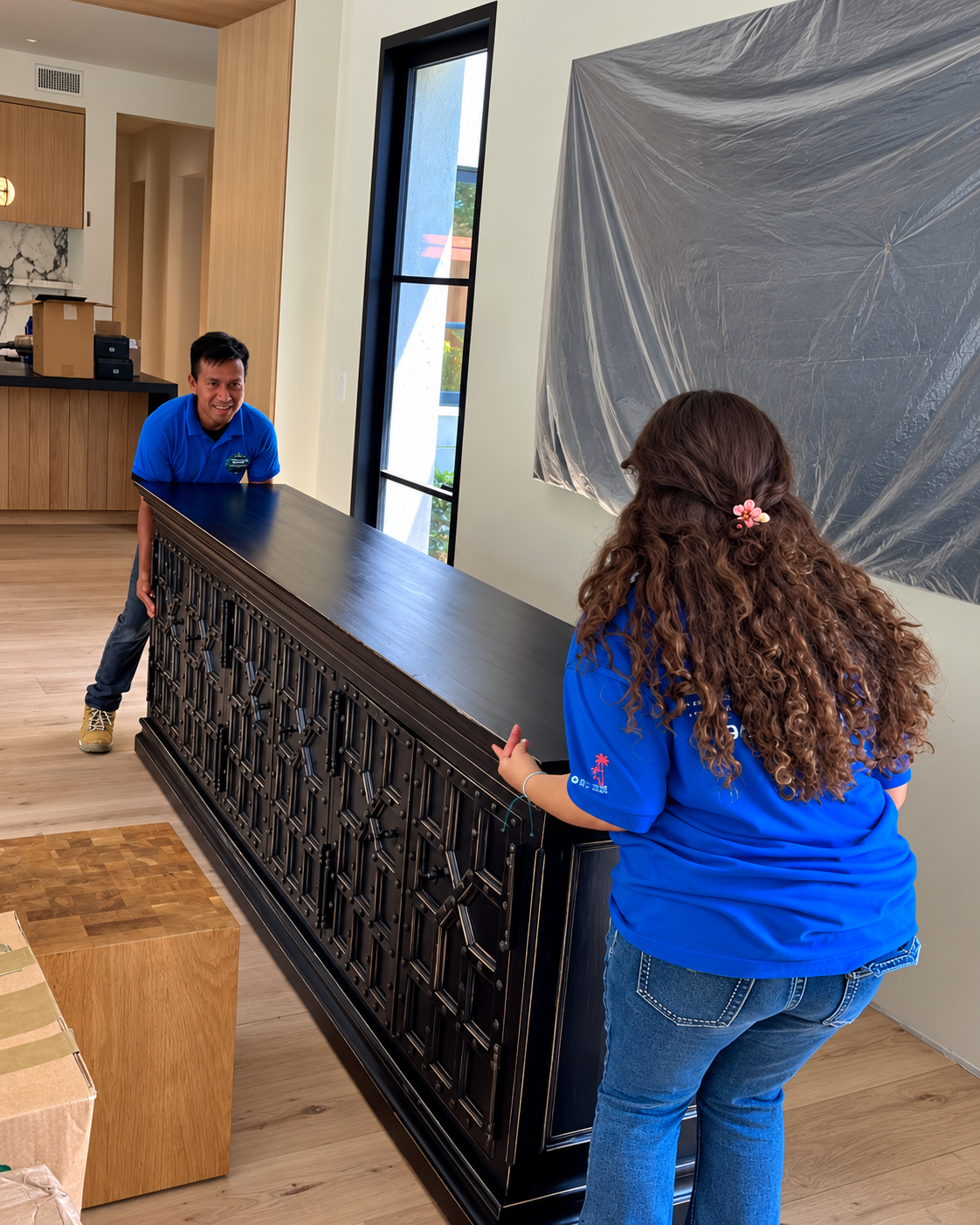 Two movers in royal-blue polos and jeans carrying a long black sideboard through a bright living room