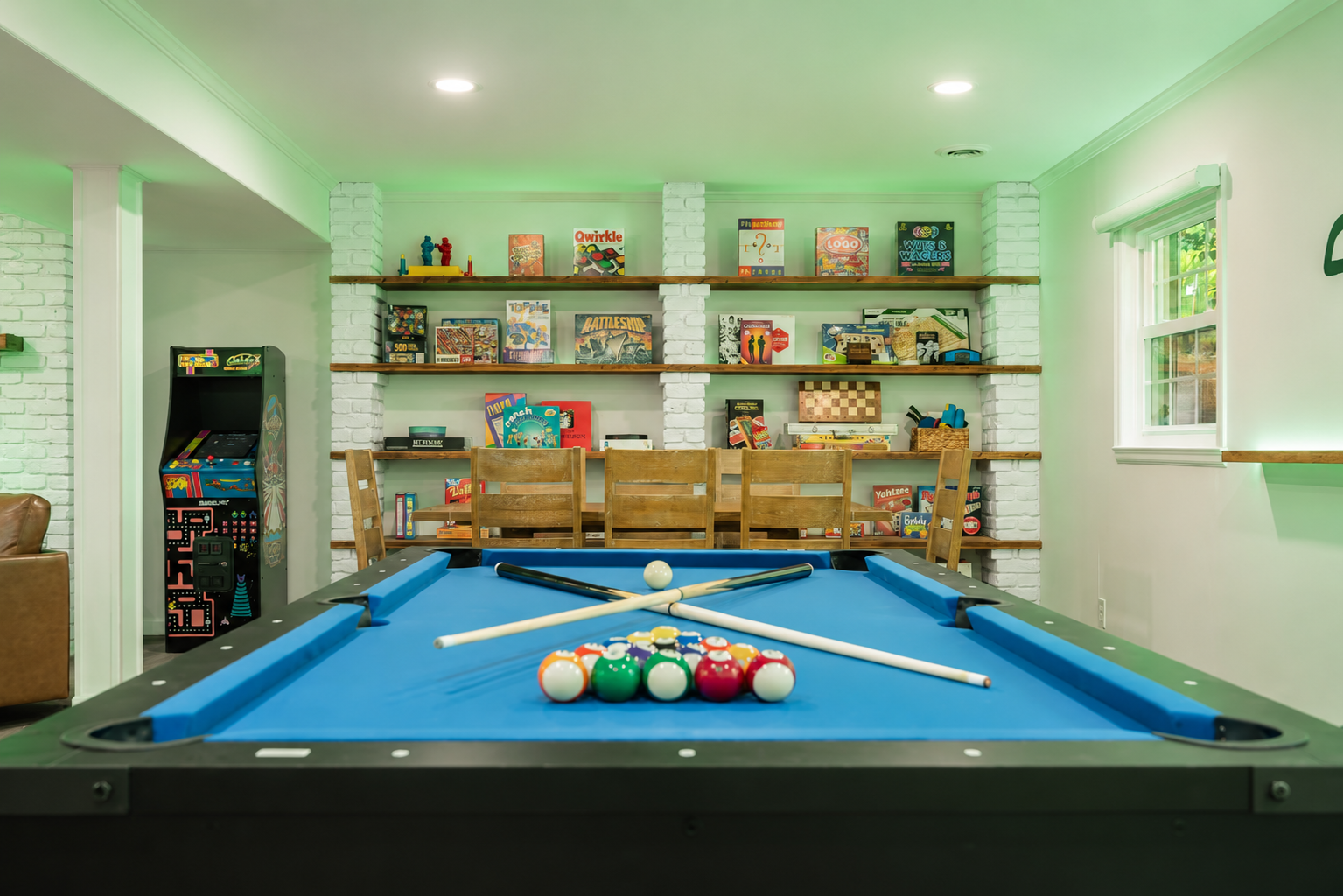 Short-term rental game room with blue pool table, arcade machine, board games, built-in shelving, and group seating. Short-term rental game room with blue pool table, arcade machine, board games, built-in shelving, and group seating.