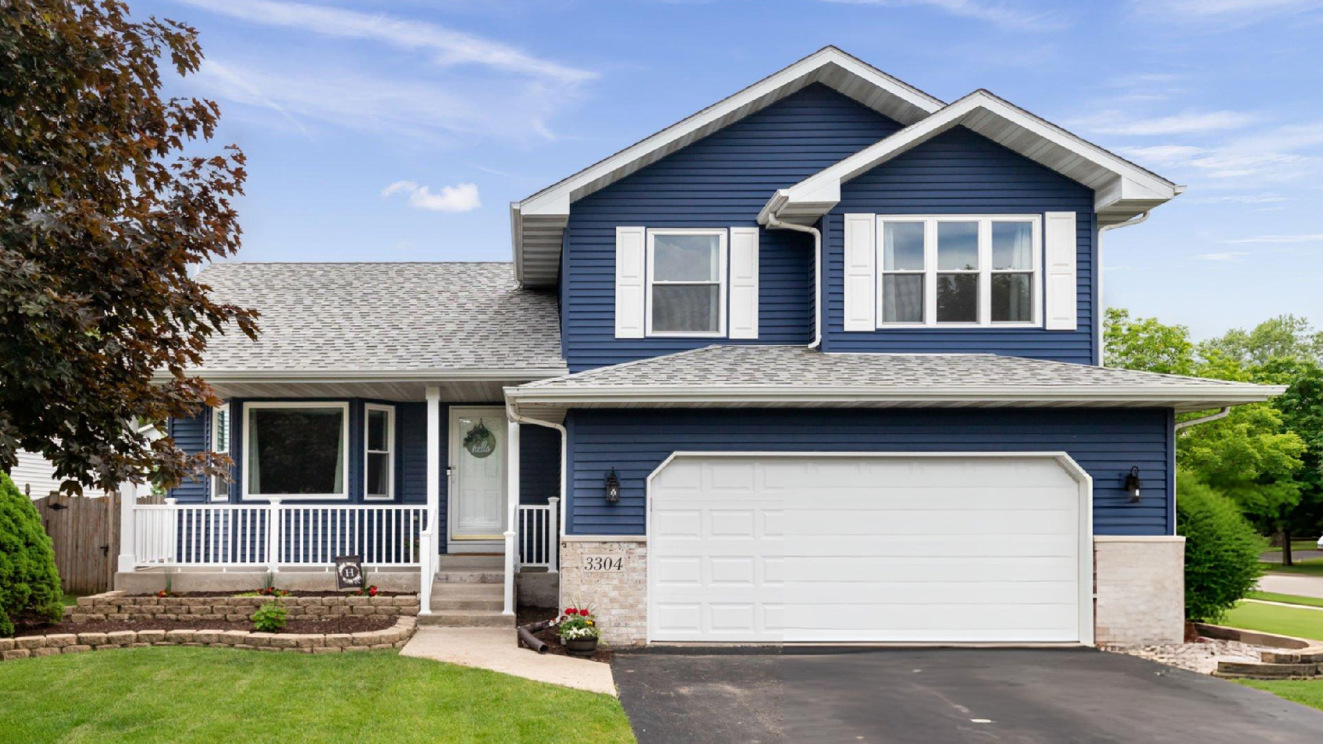 Blue suburban family home with white trim, landscaped lawn, driveway, garage, and clear sunny sky outside.