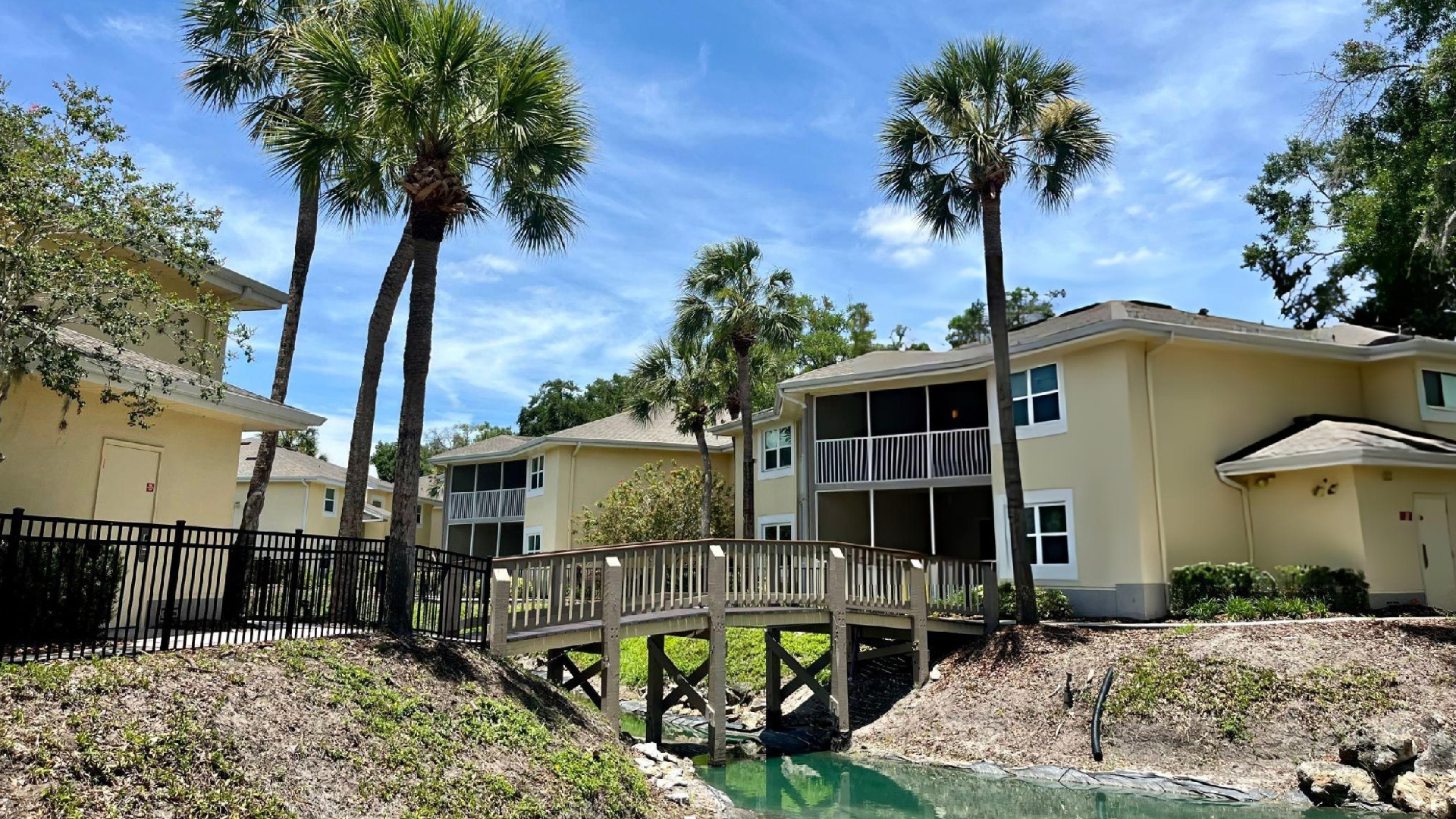 Yellow residential buildings surrounded by tall palm trees and connected by a wooden footbridge over water.