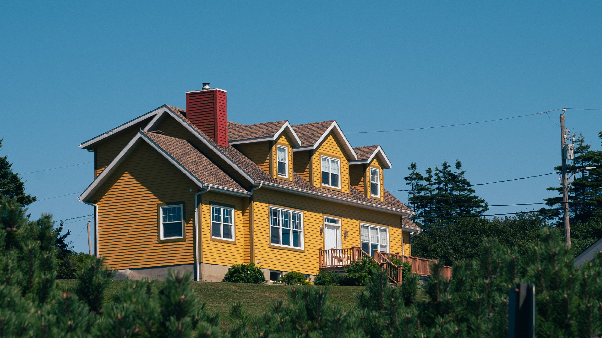 Yellow two-story house with dormer windows, red chimney, wooden siding, trees, and clear blue sky above. Yellow two-story house with dormer windows, red chimney, wooden siding, trees, and clear blue sky above.