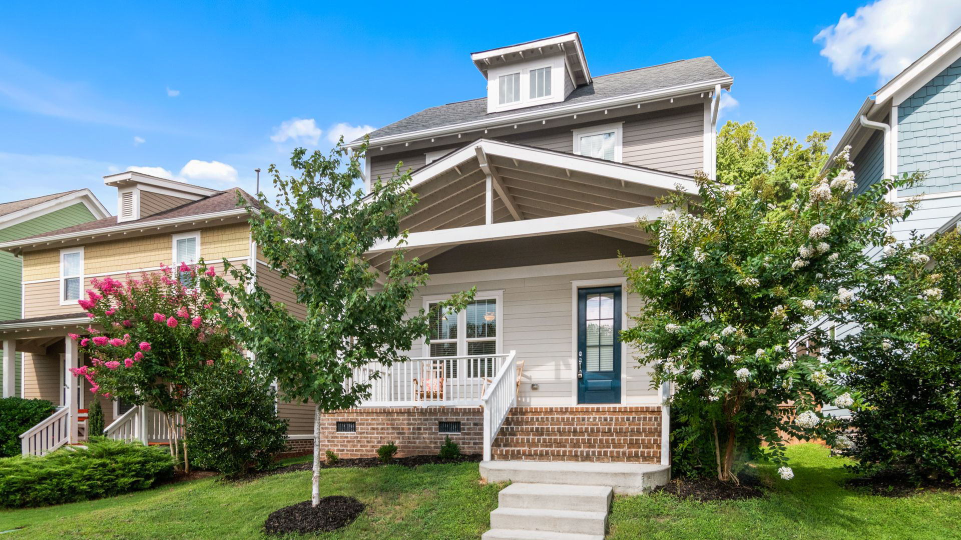 Charming suburban home with covered porch, brick steps, flowering trees, green lawn, and bright blue sky. Charming suburban home with covered porch, brick steps, flowering trees, green lawn, and bright blue sky.