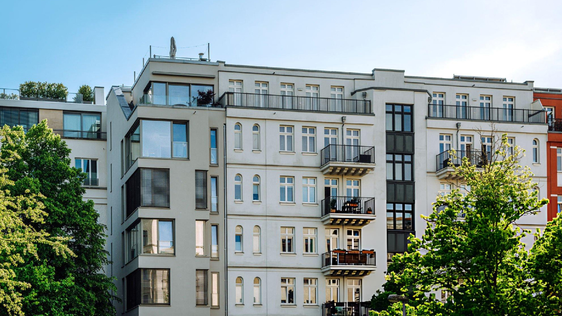 White apartment building facade with black-framed windows, balconies, surrounded by green trees under clear blue sky. White apartment building facade with black-framed windows, balconies, surrounded by green trees under clear blue sky.