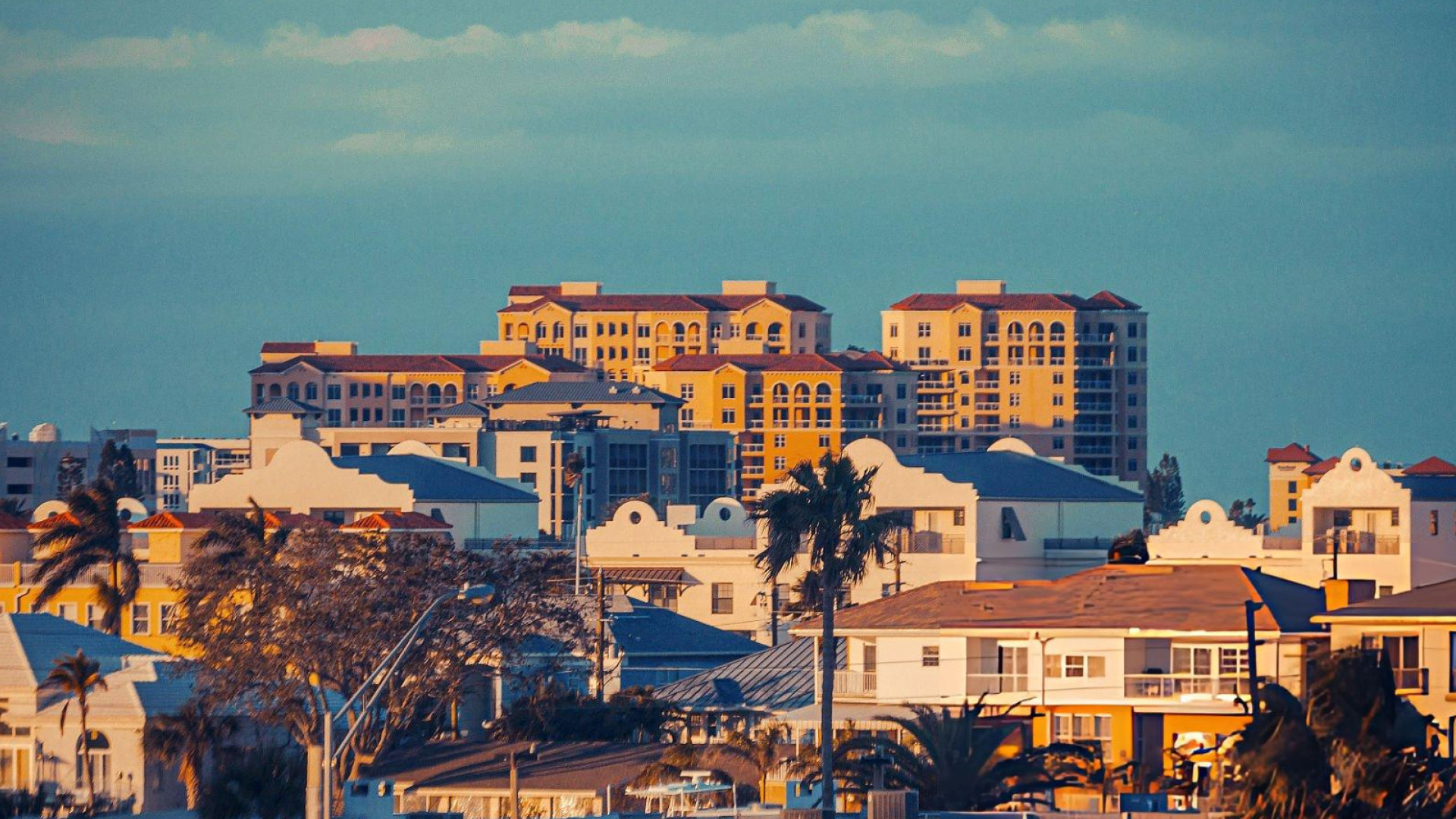 Golden evening light on yellow high-rise condos and low coastal homes with palm trees beneath a teal sky.