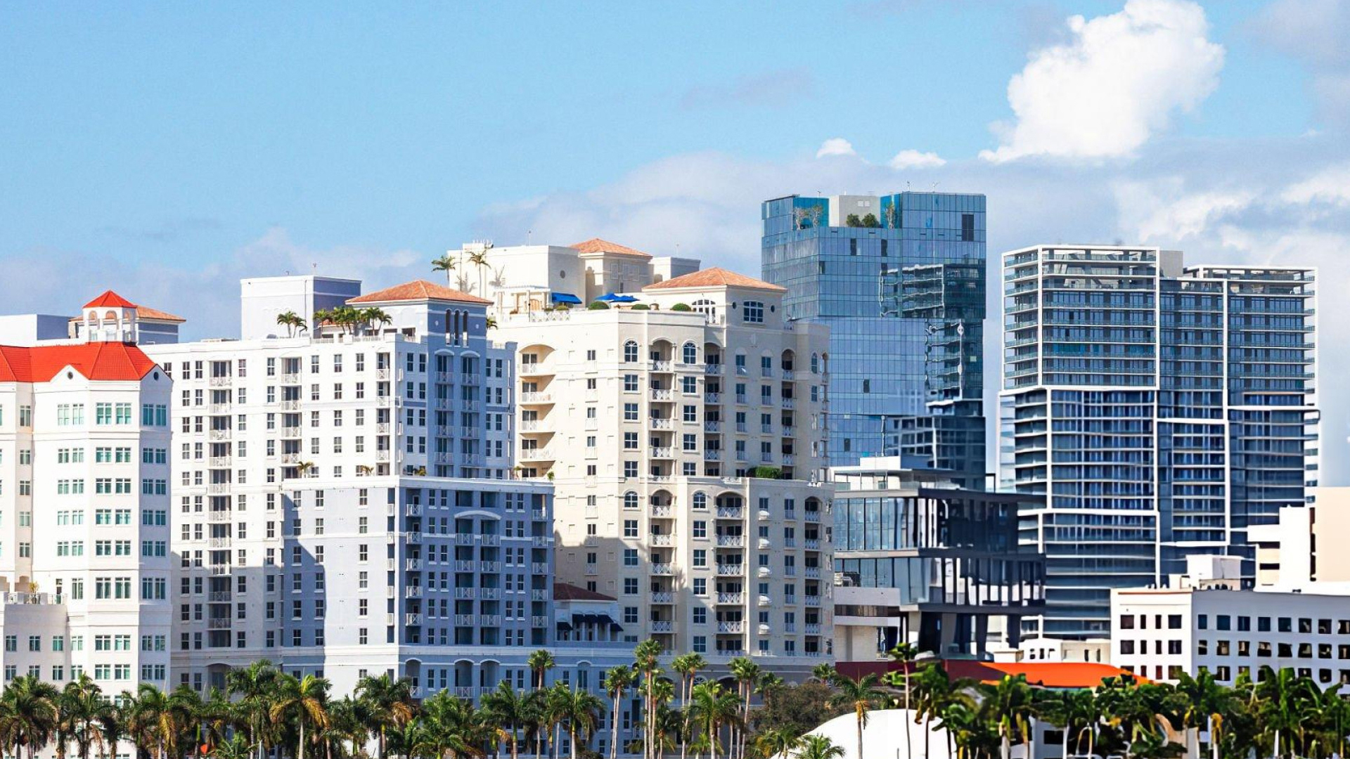 Dense city skyline of white and glass high-rise condo buildings surrounded by palm trees on a sunny day.