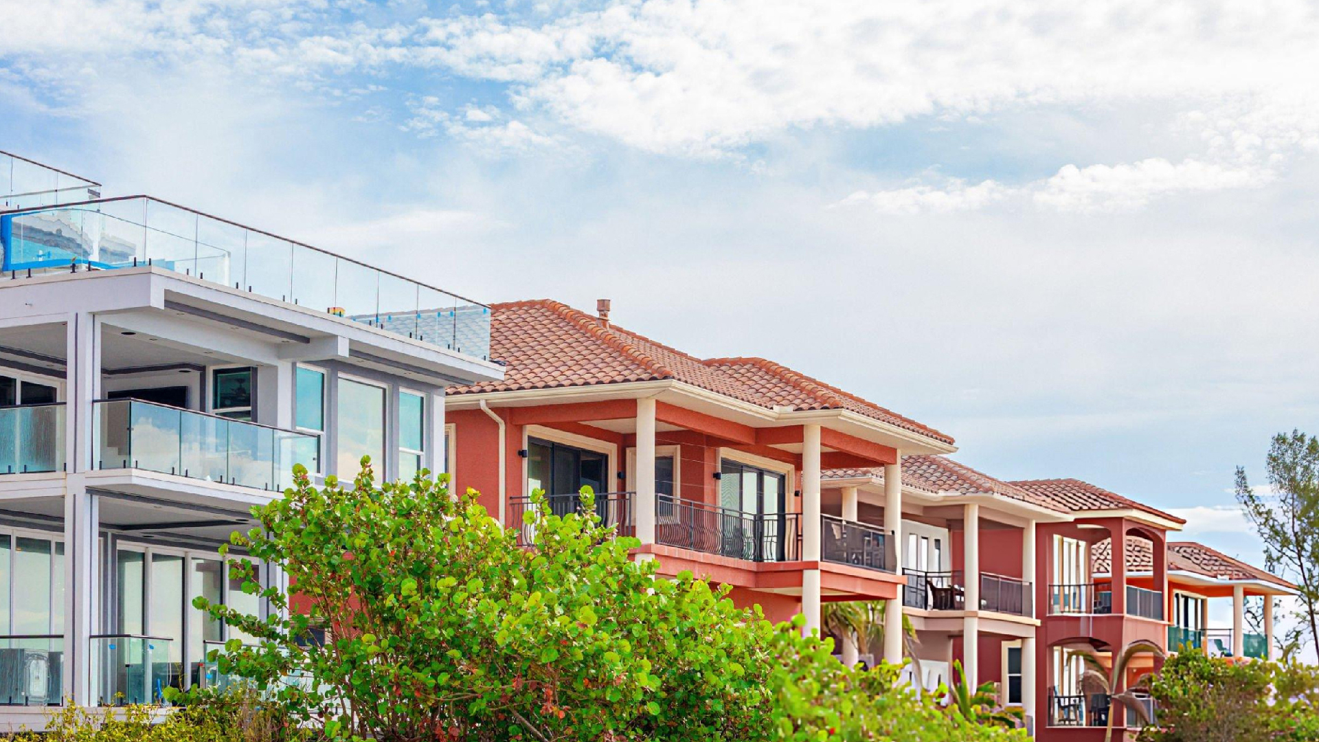 Colorful beachfront homes with red tile roofs, glass balconies, and green shrubs under a bright, partly cloudy sky.