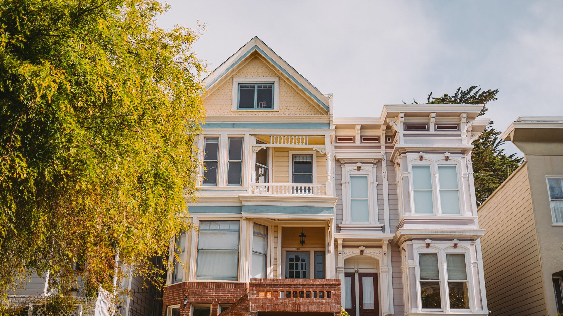 Colorful Victorian houses in yellow and lavender with brick steps, framed by a sunlit tree under cloudy sky. Colorful Victorian houses in yellow and lavender with brick steps, framed by a sunlit tree under cloudy sky.