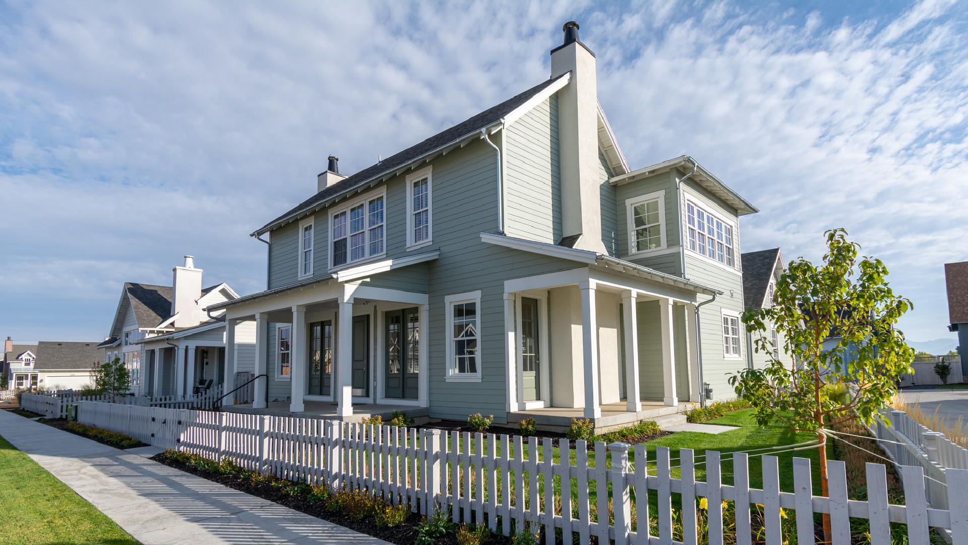Sage green two-story house with white porch and picket fence under a bright, cloudy blue sky. Sage green two-story house with white porch and picket fence under a bright, cloudy blue sky.